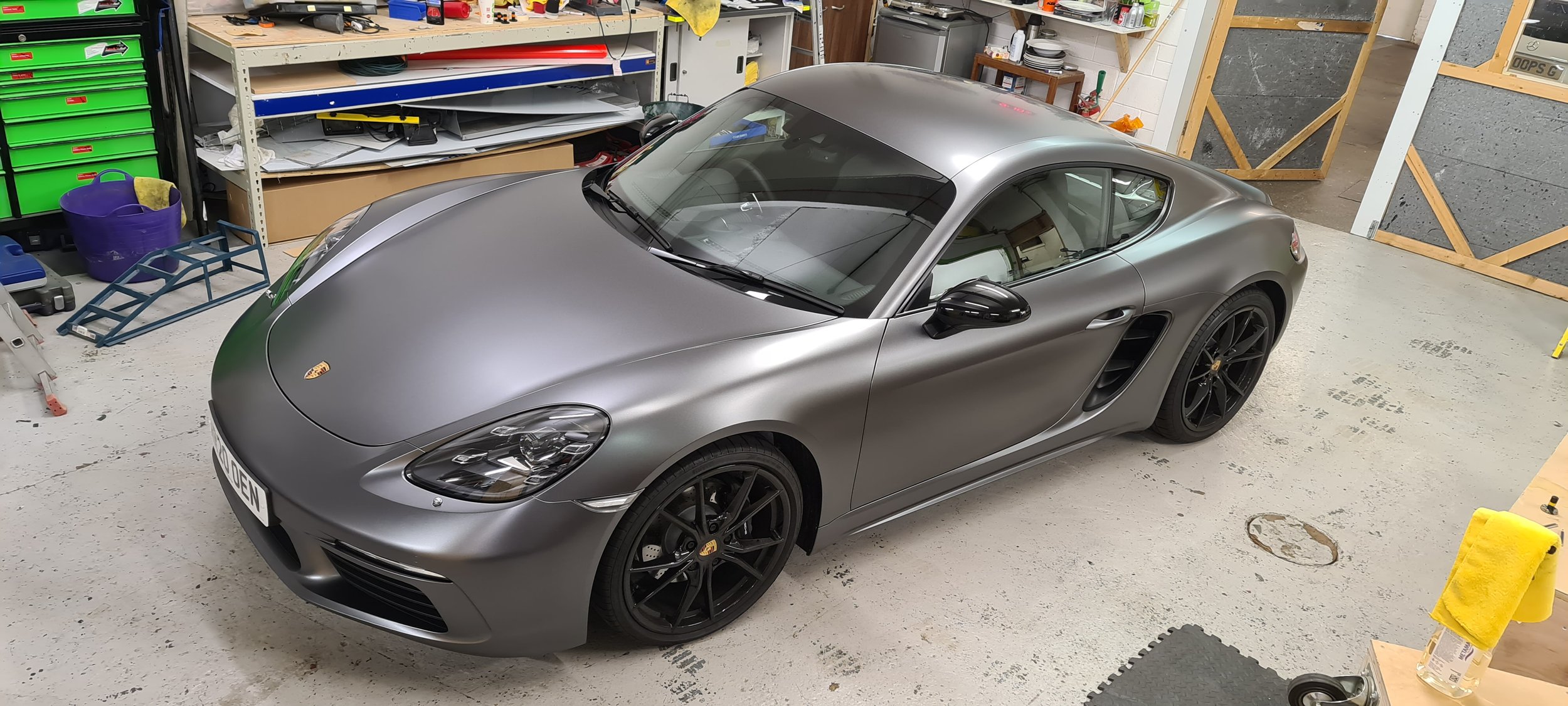 A metallic grey Porsche sports car parked inside a cluttered garage with tools and storage shelves.