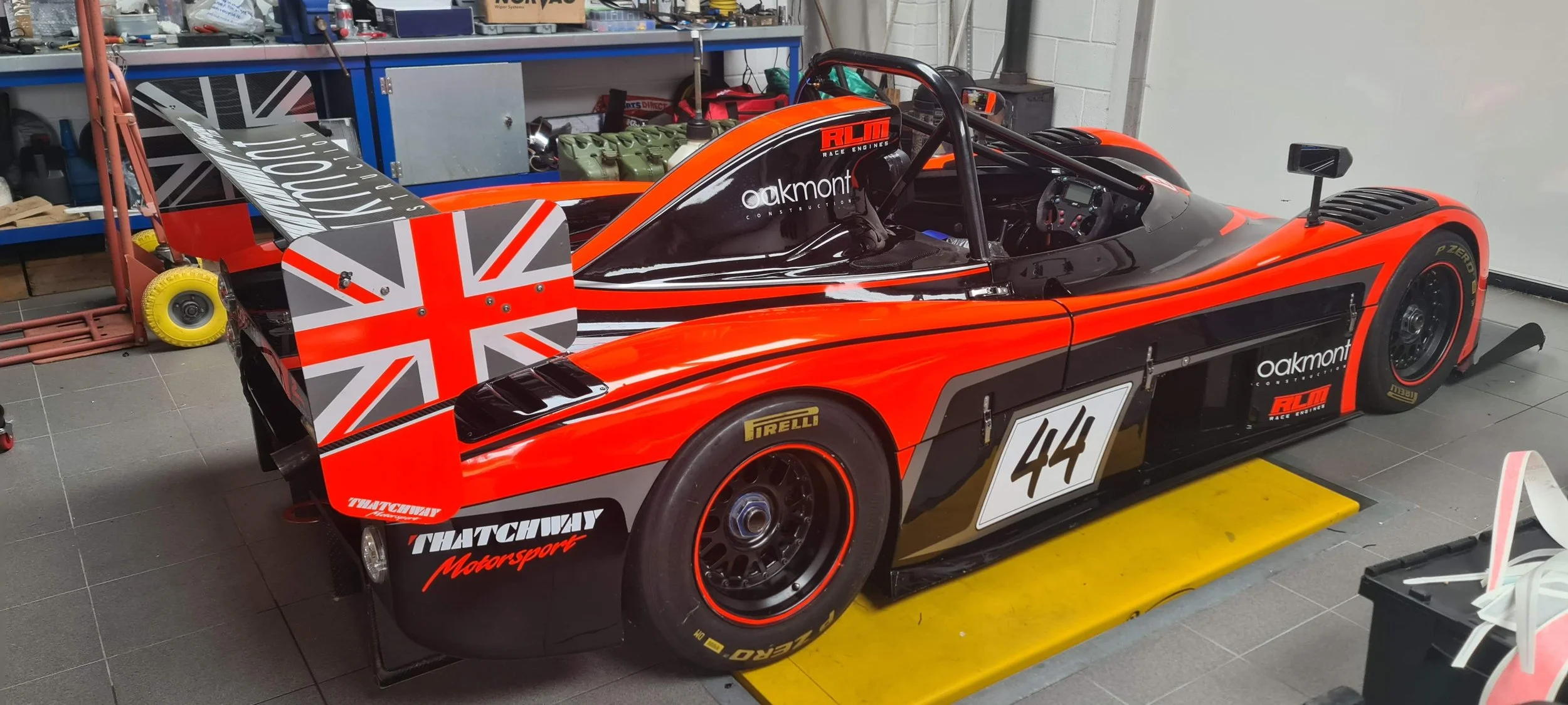 A red and black race car with various sponsor logos, number 44, inside a garage workshop with tools and equipment in the background.