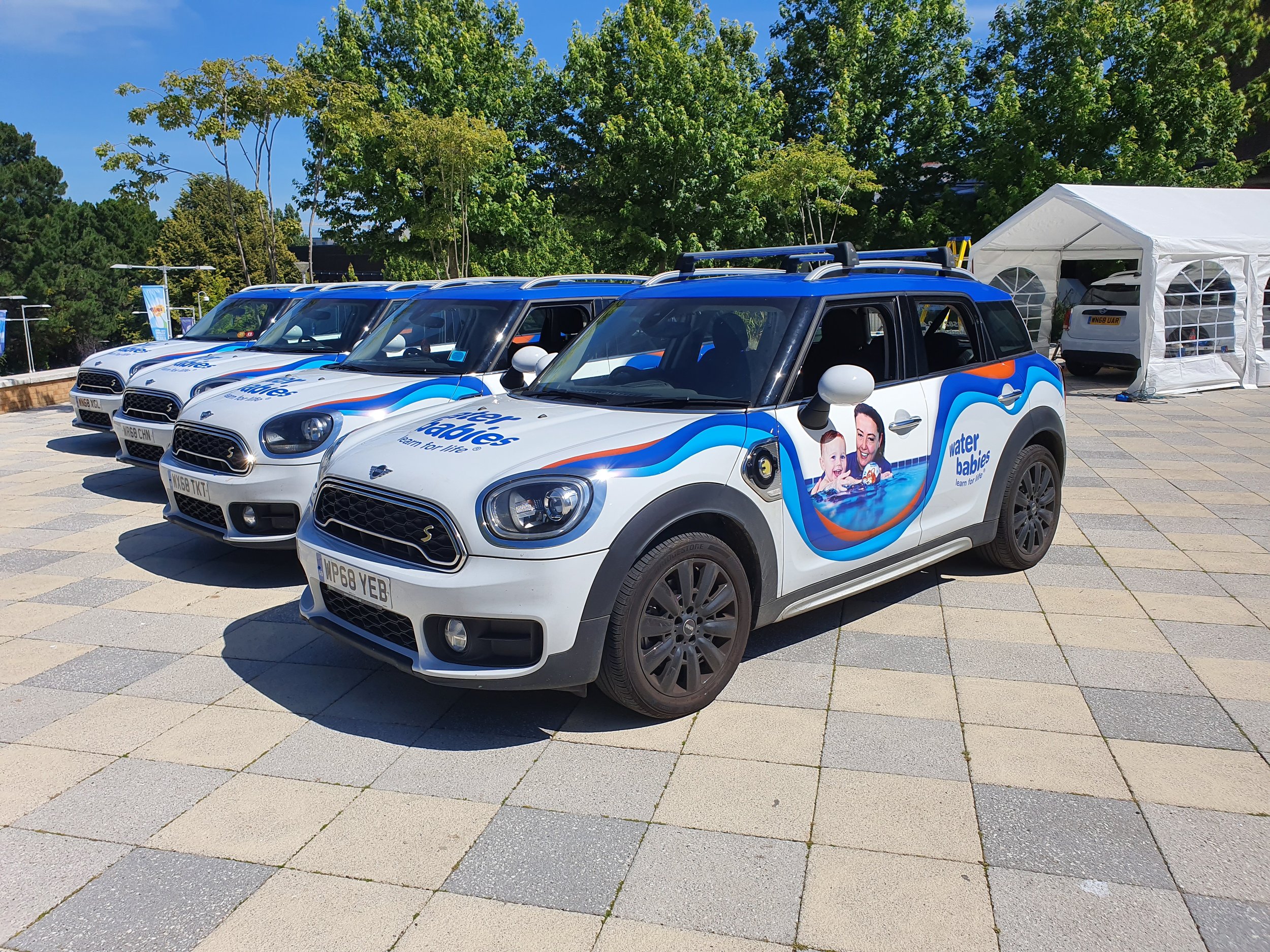 Multiple white electric cars with blue and orange branding for 'Water Babies' parked in a row outdoors on a paved surface, with a large green tree and a white tent in the background.