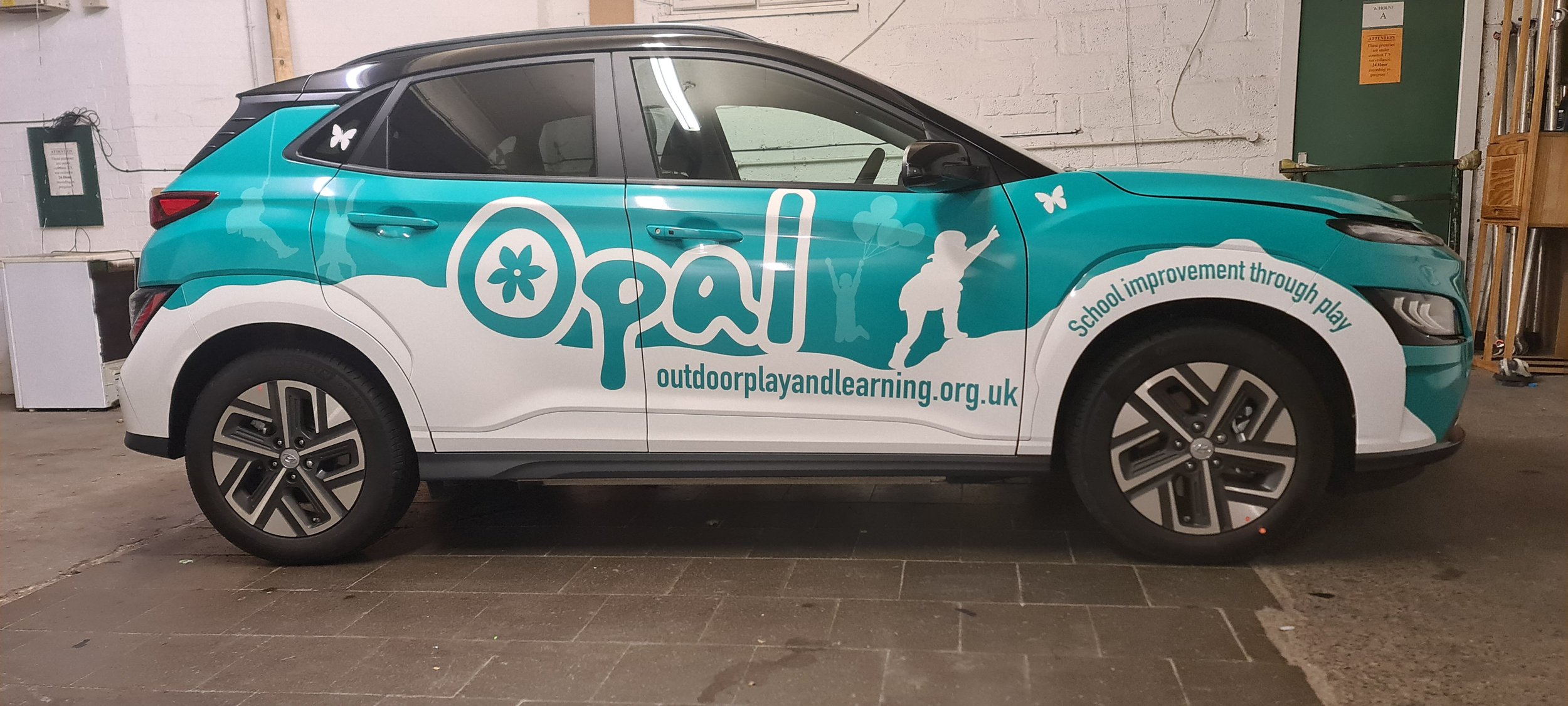 A teal and white vehicle with the word 'Opa' and various child-related graphics and text promoting outdoor play and learning for school improvement, parked indoors.