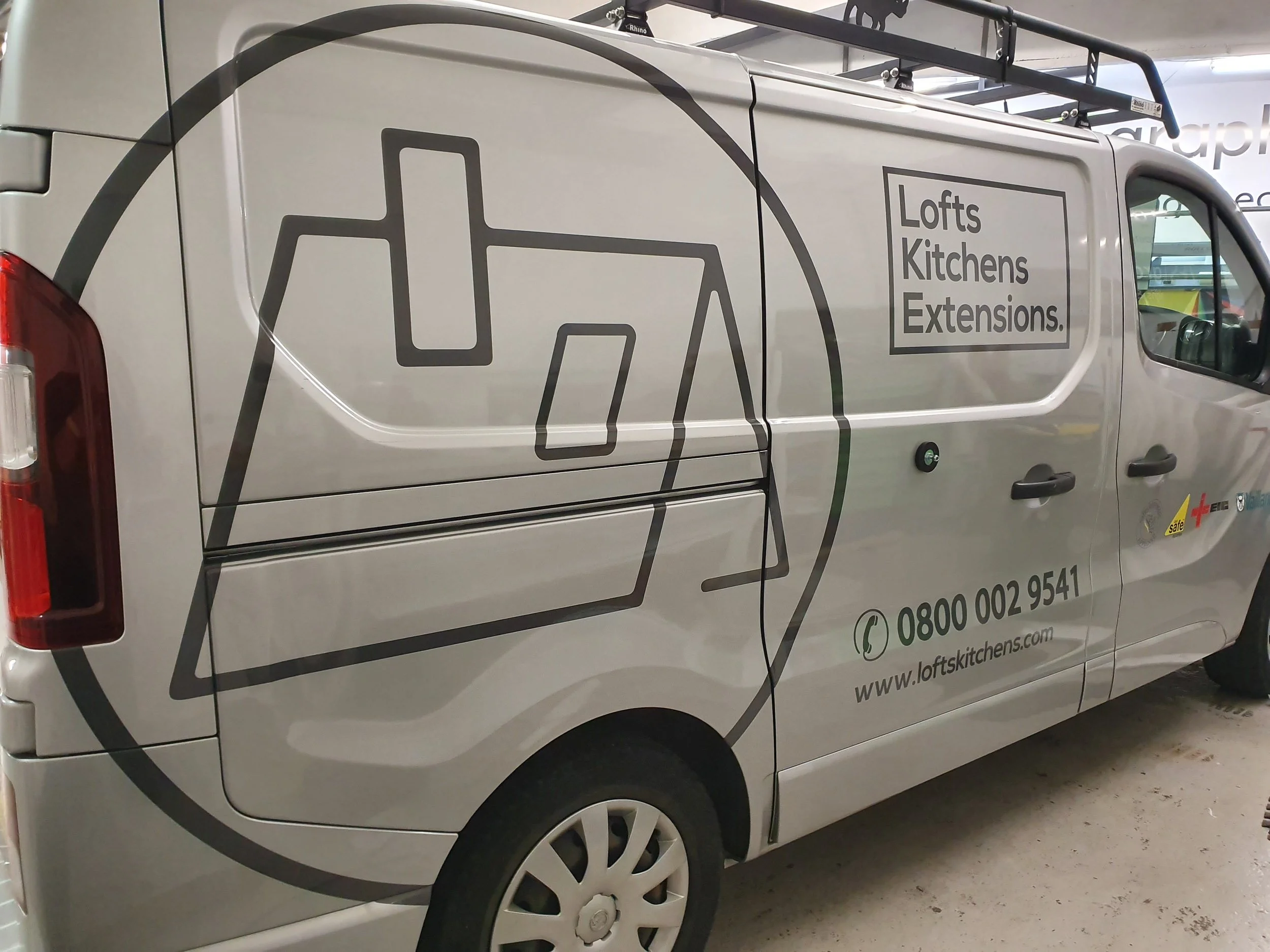 A white van with black line art graphics and text promoting Lofts, Kitchens, Extensions. The graphics include a sketch of a chair and various rectangular shapes. Contact information and a website URL are visible on the side of the van.