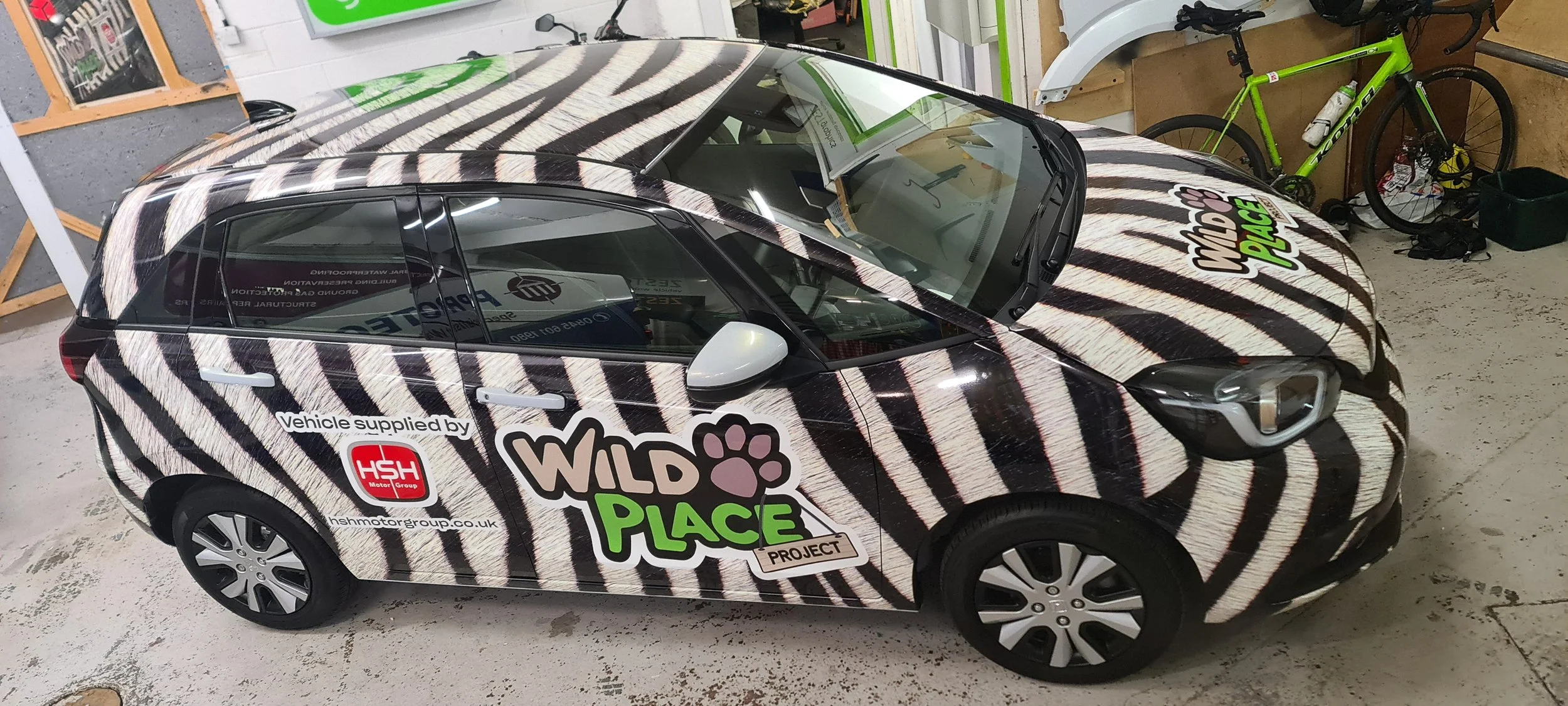 A small car with a zebra print wrap featuring the logos and text for the Wild Place Project, parked indoors next to bicycles and other items.