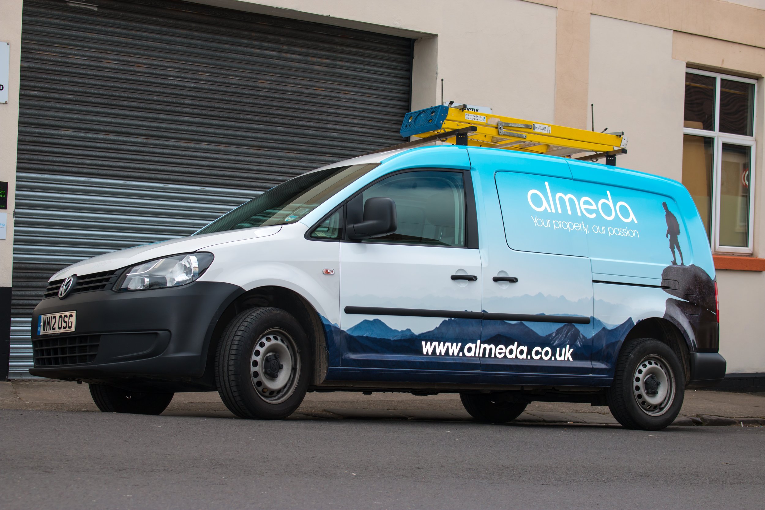 A white and blue delivery van with the logo and website of 'Almeda' parked on the street in front of a building with a metal roll-up door and windows. The van has a ladder mounted on top.