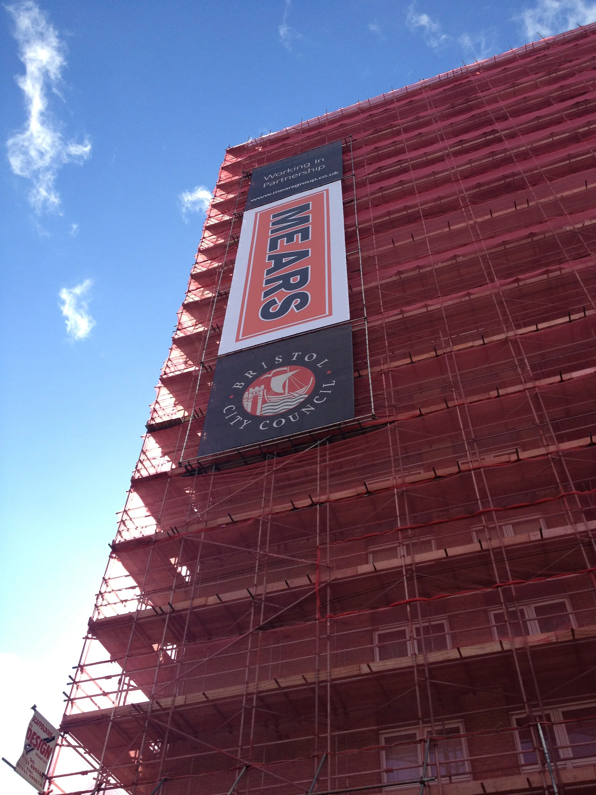 A tall building under construction covered with red scaffolding. There is a large vertical banner that reads 'WE ARE' with a logo beneath, and another banner at the top that says 'Working in Partnership' with a URL.