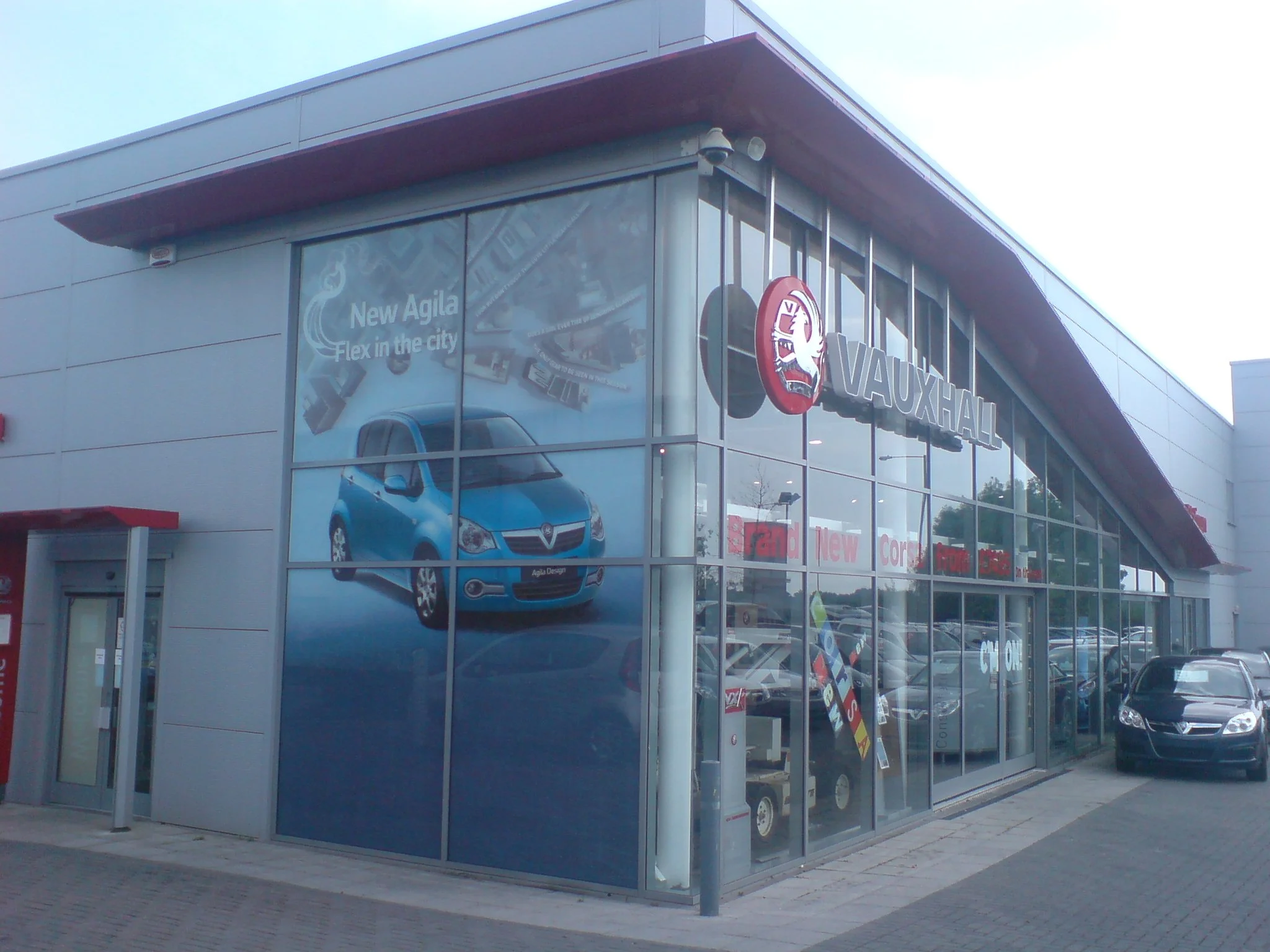 Exterior view of a modern Vauxhall car dealership with large glass windows displaying a blue car inside, promotional posters on the glass, and a parking lot with a few cars outside.
