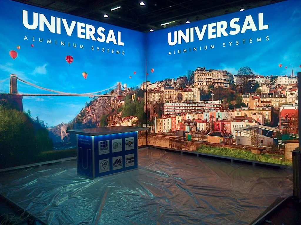 Trade show booth for Universal Aluminium Systems with a backdrop of a cityscape including a bridge and hot air balloons in the sky.