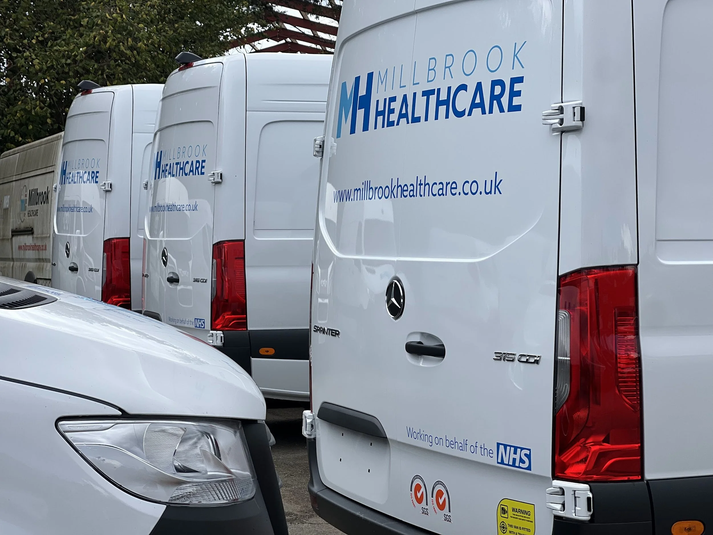 A row of white healthcare vans with vehicle graphics for Millbrook Healthcare and NHS on the side, parked closely together, with a silver car in front.