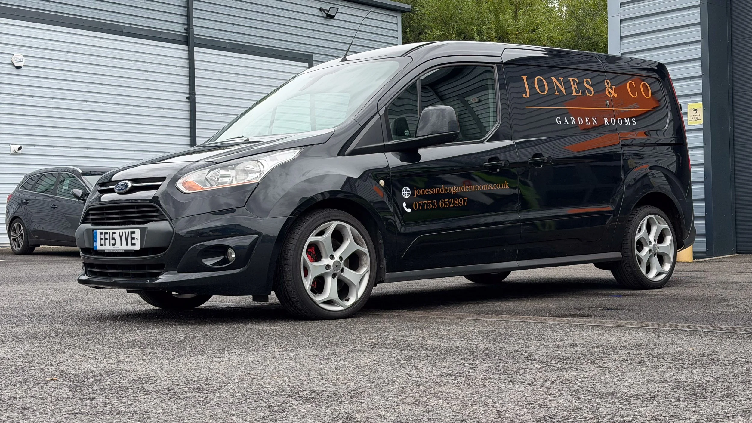 A black Ford Transit van parked next to a gray car near a modern industrial building. The van has orange and white text on its side advertising Jones & Co Garden Rooms, including a website and phone number.