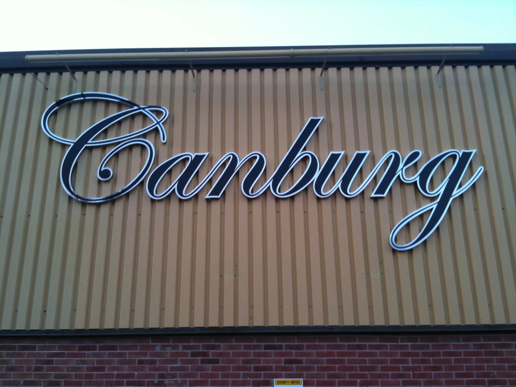 Close-up of a store or building exterior with beige vertical siding, a black neon sign reading 'Camehanging' in cursive font, and a brick wall at the bottom with a small yellow security notice sign.