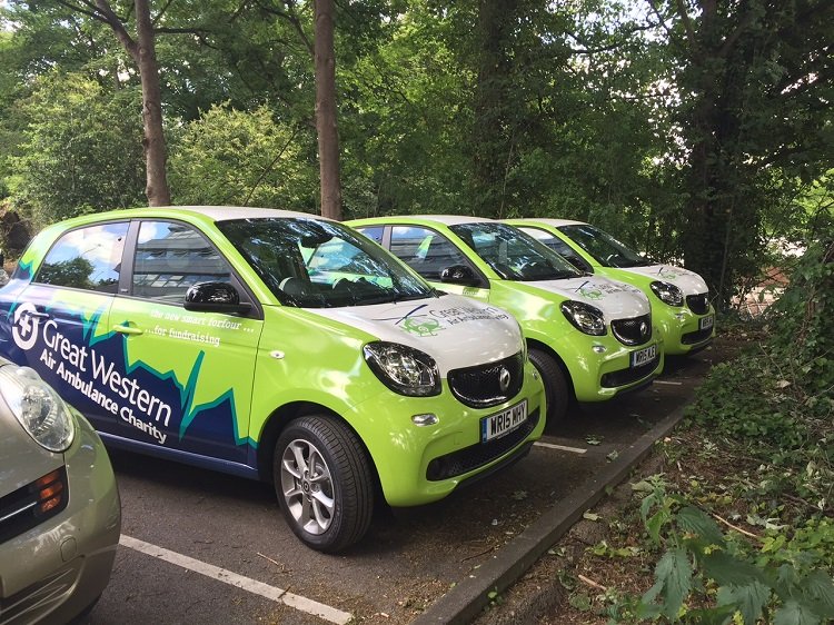 Four green vehicles parked in a row on a parking lot next to trees, with the side of a gray car partially visible in the foreground. The vehicles are branded with the Great Western Air Ambulance Charity logo.