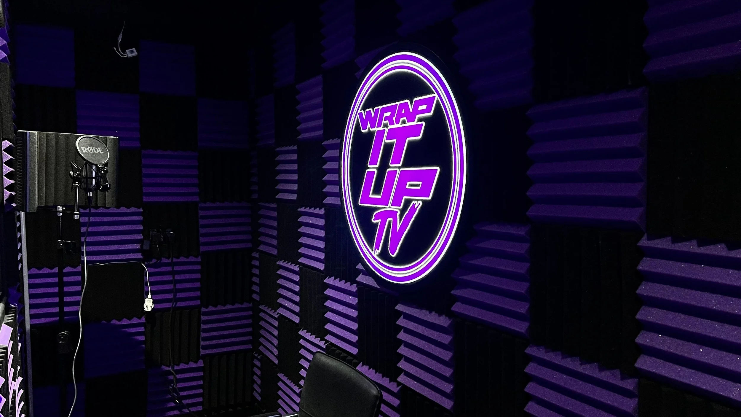Recording studio with purple acoustic foam panels, a neon sign reading "Wrap It Up TV," and recording equipment including a microphone on a stand.