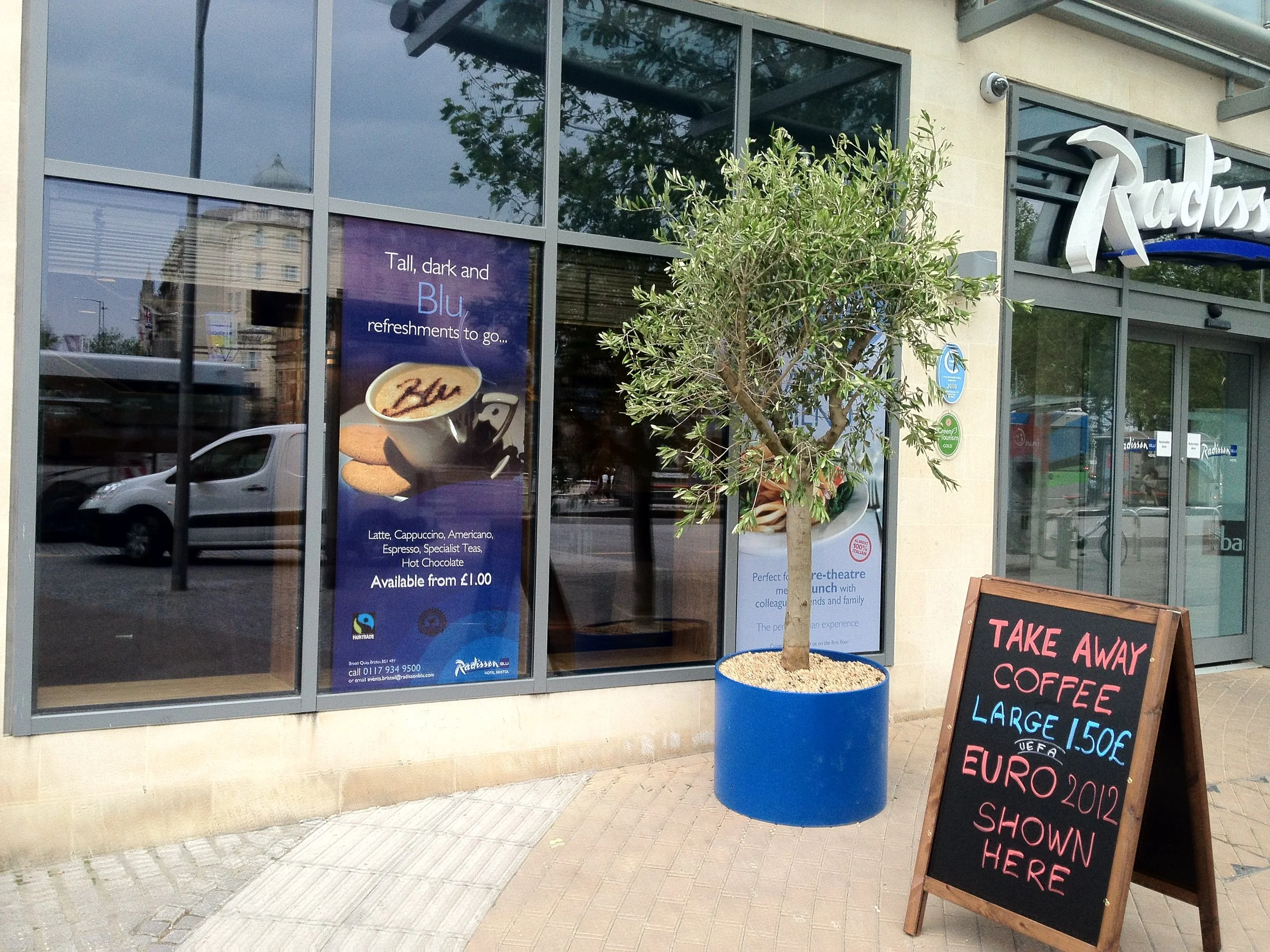 The image shows the exterior of a cafe with large windows, a potted tree in a blue container, and a blackboard sign advertising take-away coffee. A promotional poster on the window features a coffee cup and cookies, with text about coffee options and