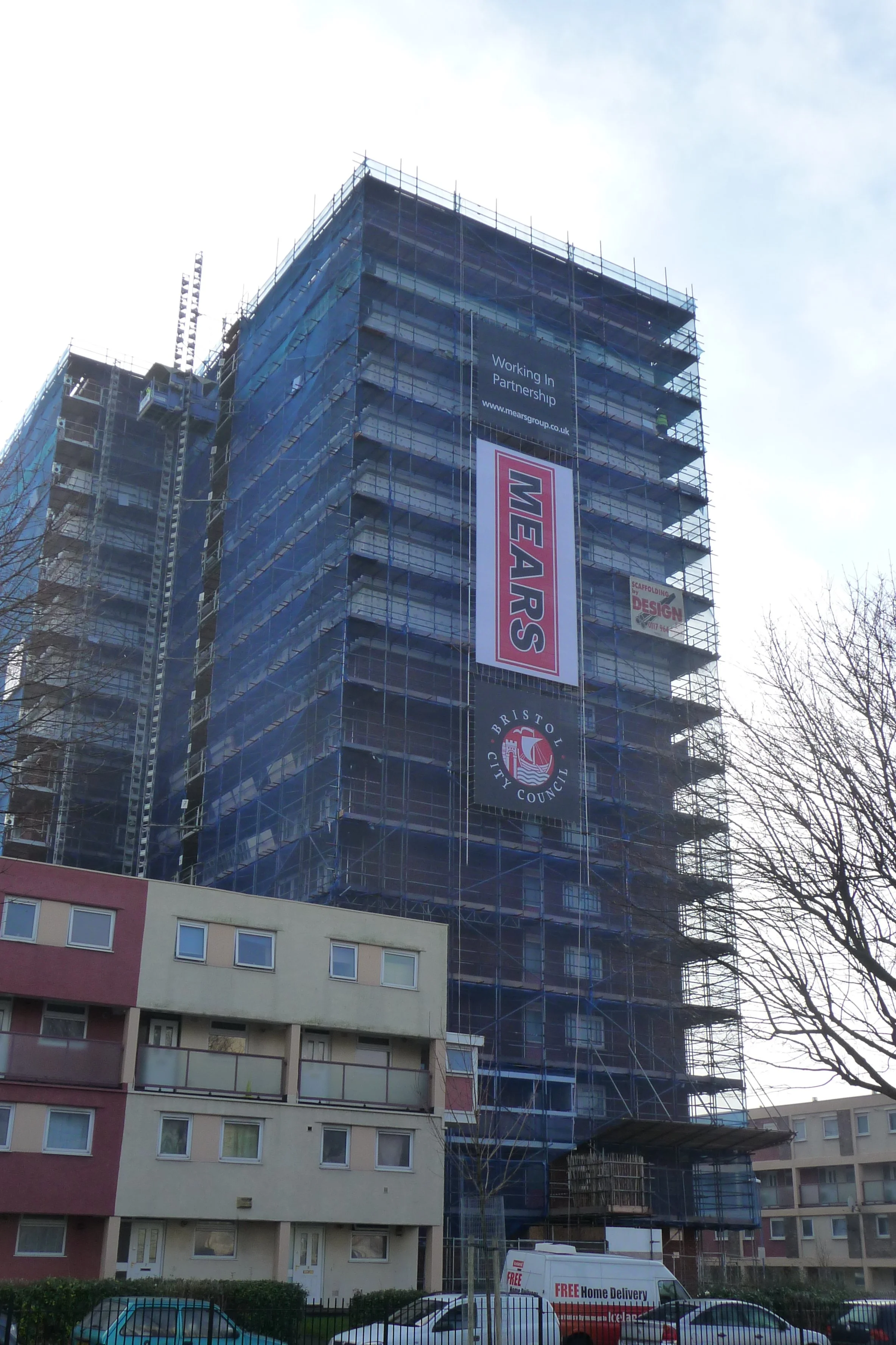 A tall building under construction with scaffolding around it. The building has signs that say "Working in Partnership" and "Wear S". In front of the building, there are parked cars and a delivery van, with residential buildings behind a fence.