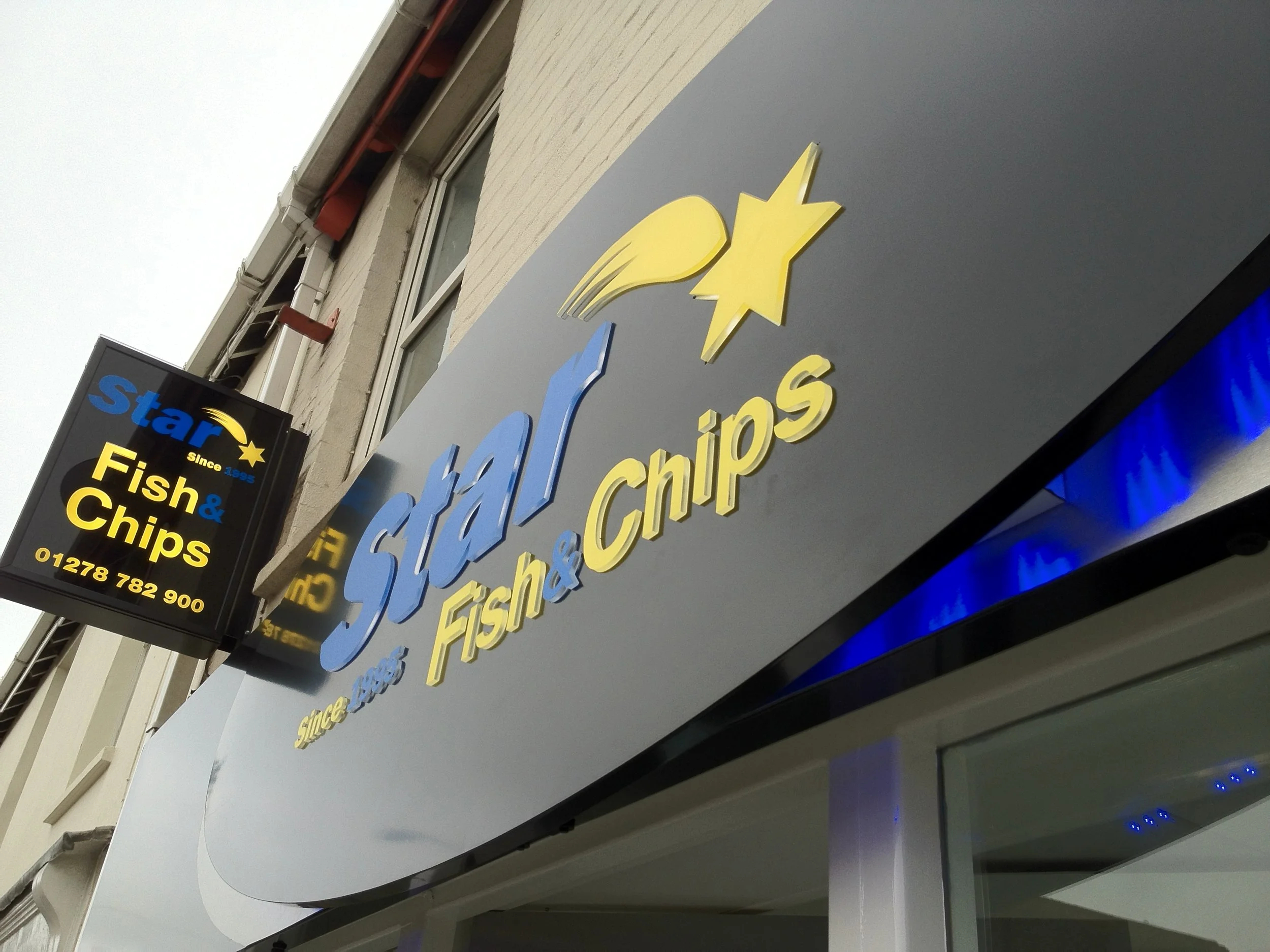 A storefront sign for Star Fish & Chips, with yellow and blue lettering and a star logo, mounted on a building with a beige brick exterior and attached awning.