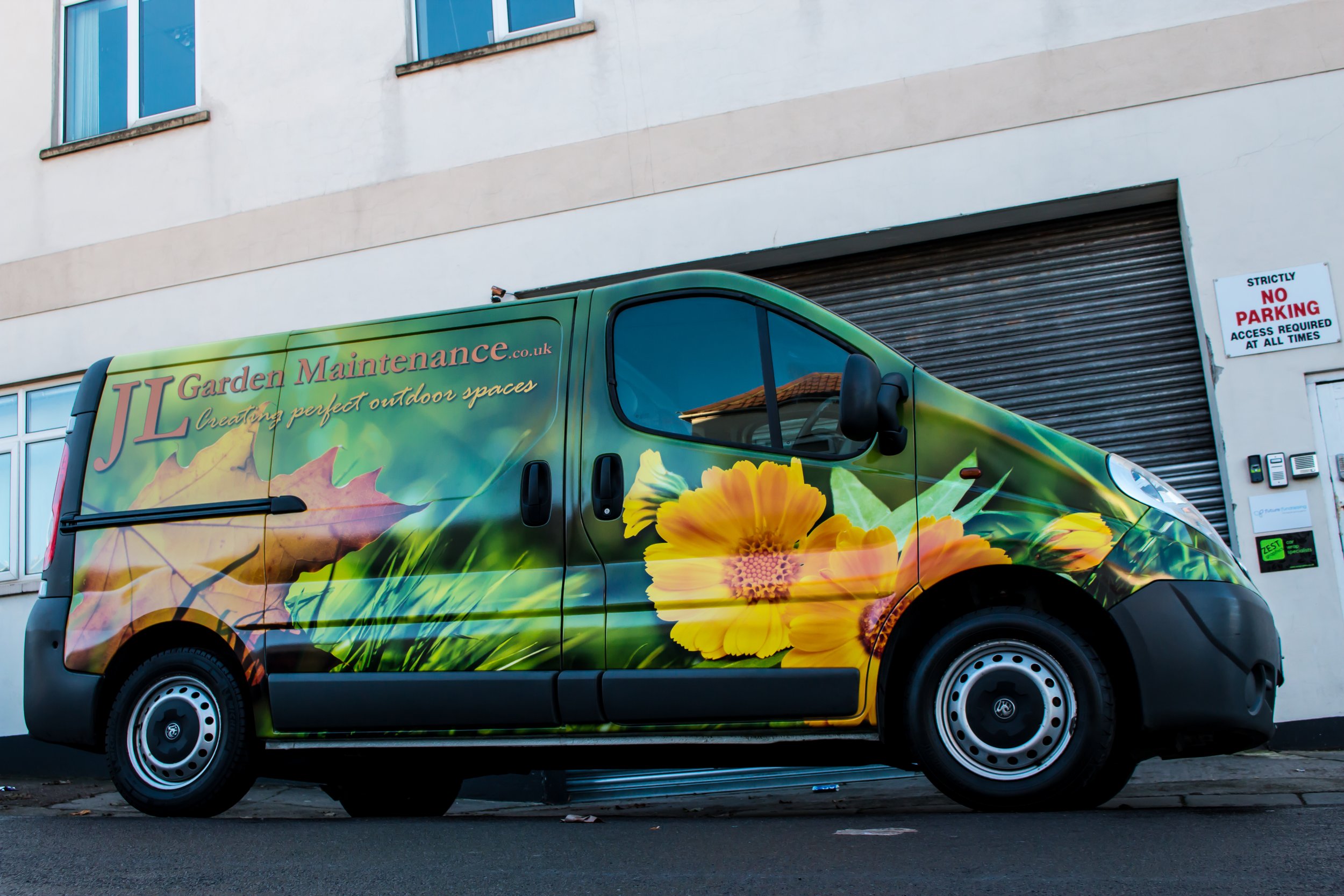 A delivery van with a vibrant floral and greenery wrap, parked in front of a building with a 'No Parking' sign and closed roller shutter door.