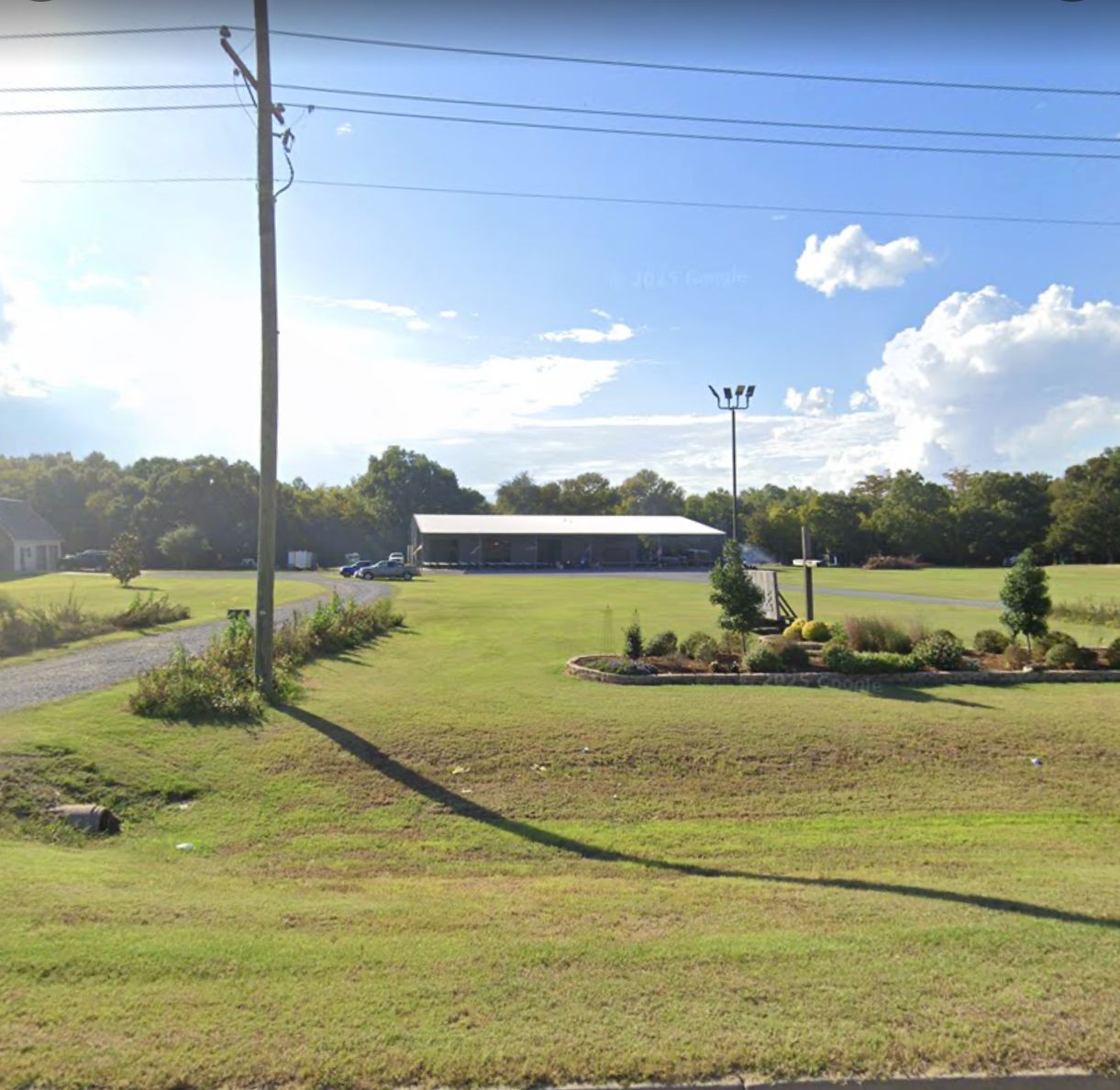 A grassy area with a few small trees and flower beds, a utility pole, a gravel pathway, and a covered building with seating in the background, under a blue sky with scattered clouds.