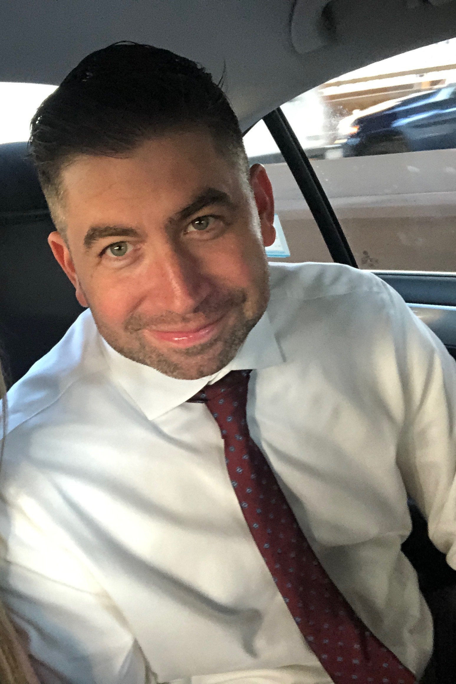 A man with dark hair, wearing a white shirt and a red patterned tie, sitting in a car with a city street visible through the window.
