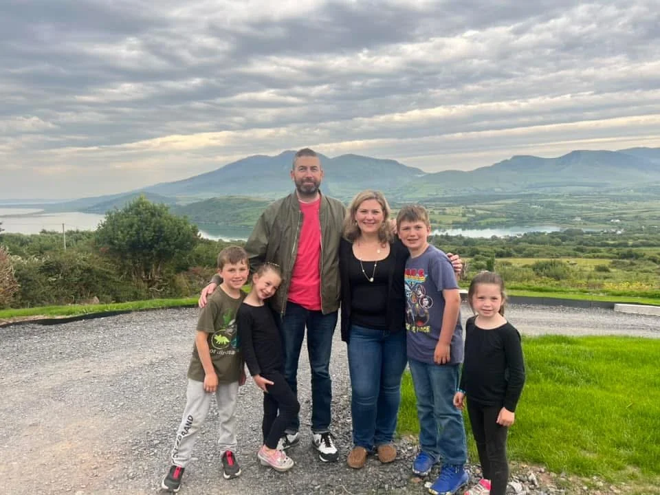 Family photo of two adults and four children standing outdoors on a gravel area, with green fields, hills, and a cloudy sky in the background.