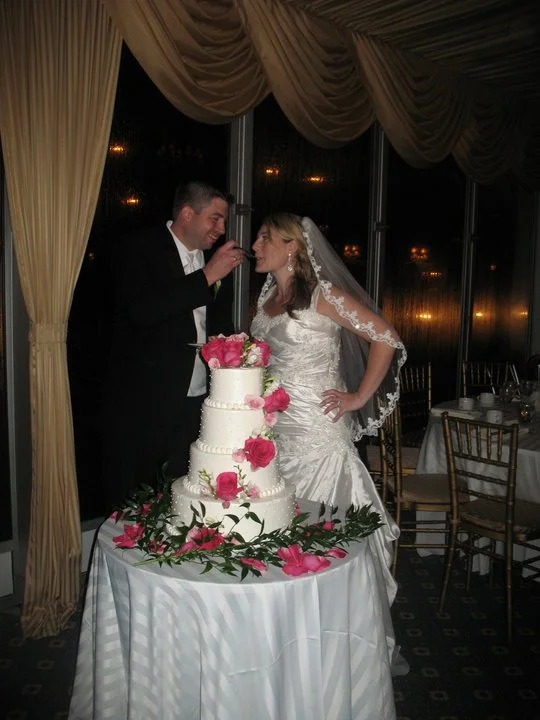 Bride and groom feeding each other cake at wedding reception, with a three-tiered wedding cake decorated with pink flowers on a table.