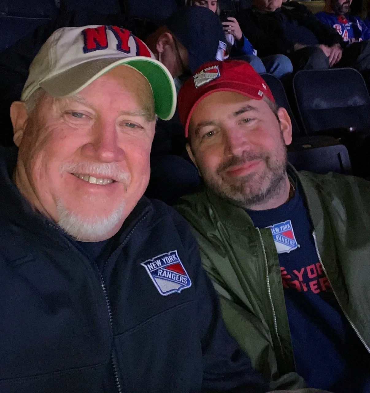 Two men smiling at the camera, seated in a stadium. They are wearing New York Rangers apparel, including jackets and hats.