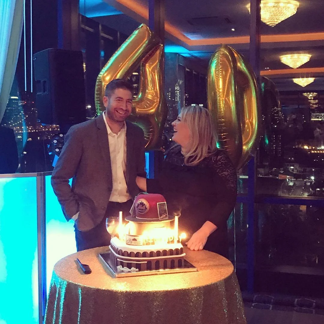 A man and woman celebrating a 40th birthday at a party with a cake, balloons, and festive decorations in a high-rise venue at night.