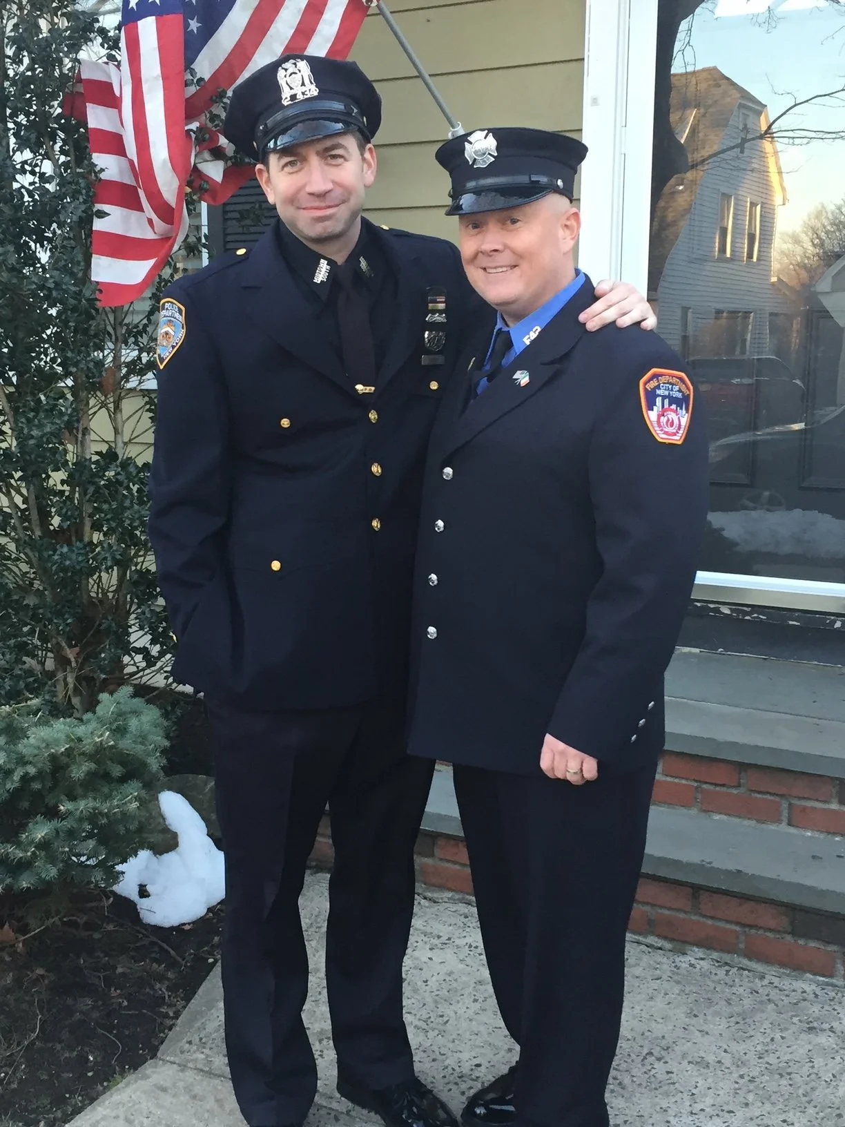 Two men in police uniforms standing outside a house, smiling, with an American flag in the background.
