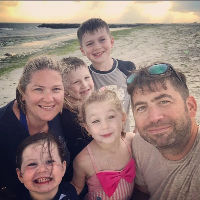 A group of six people, two adults and four children, taking a selfie on a beach during sunset, smiling and enjoying the moment.