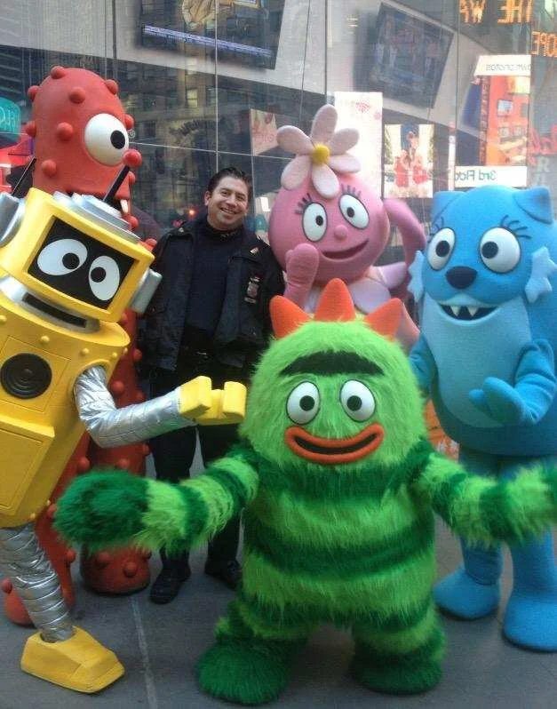 A man in a dark uniform standing in an indoor venue, surrounded by colorful mascot costumes of various characters from children's shows and cartoons, including a yellow robot, a pink flower character, a blue cat, and a green fuzzy creature, all posin