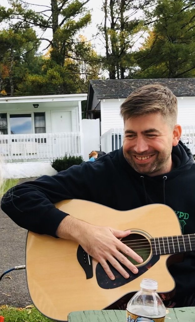 A smiling man with short brown hair and a beard playing an acoustic guitar outdoors, sitting at a table with a water bottle, with houses and trees in the background.