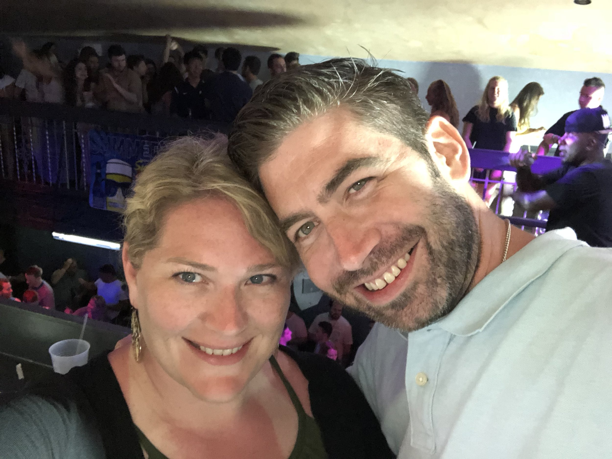 A smiling man and woman taking a selfie at a crowded indoor event with people dancing and socializing in the background.