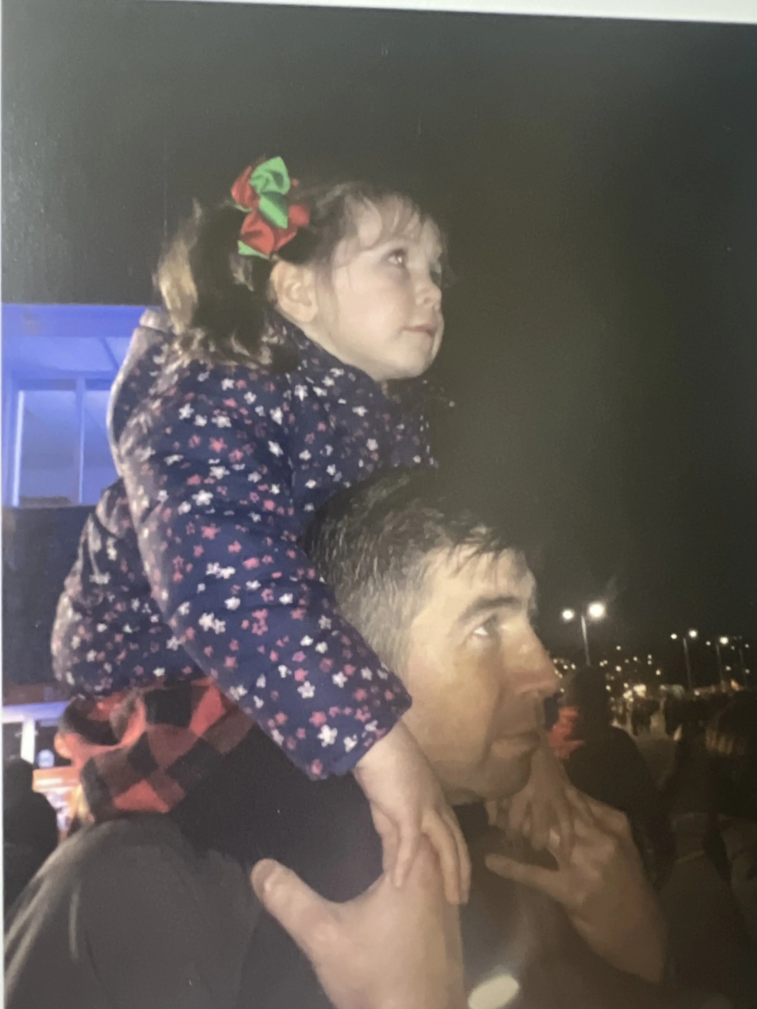 A young girl with curly hair and a rainbow-colored bow on her head sitting on the shoulders of an adult male at night. The background shows some street lights and parked cars.