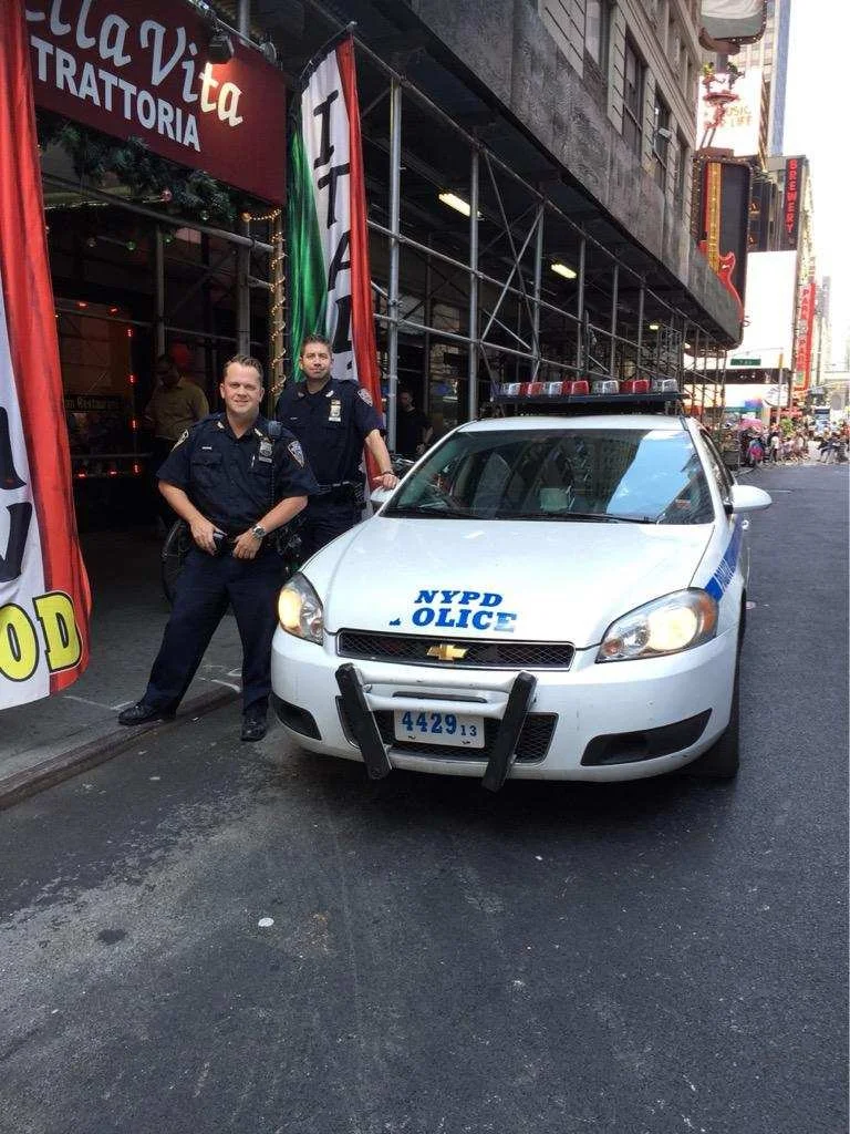 Two police officers standing next to a NYPD police car parked on a city street in front of a restaurant with red, green, and white banners.