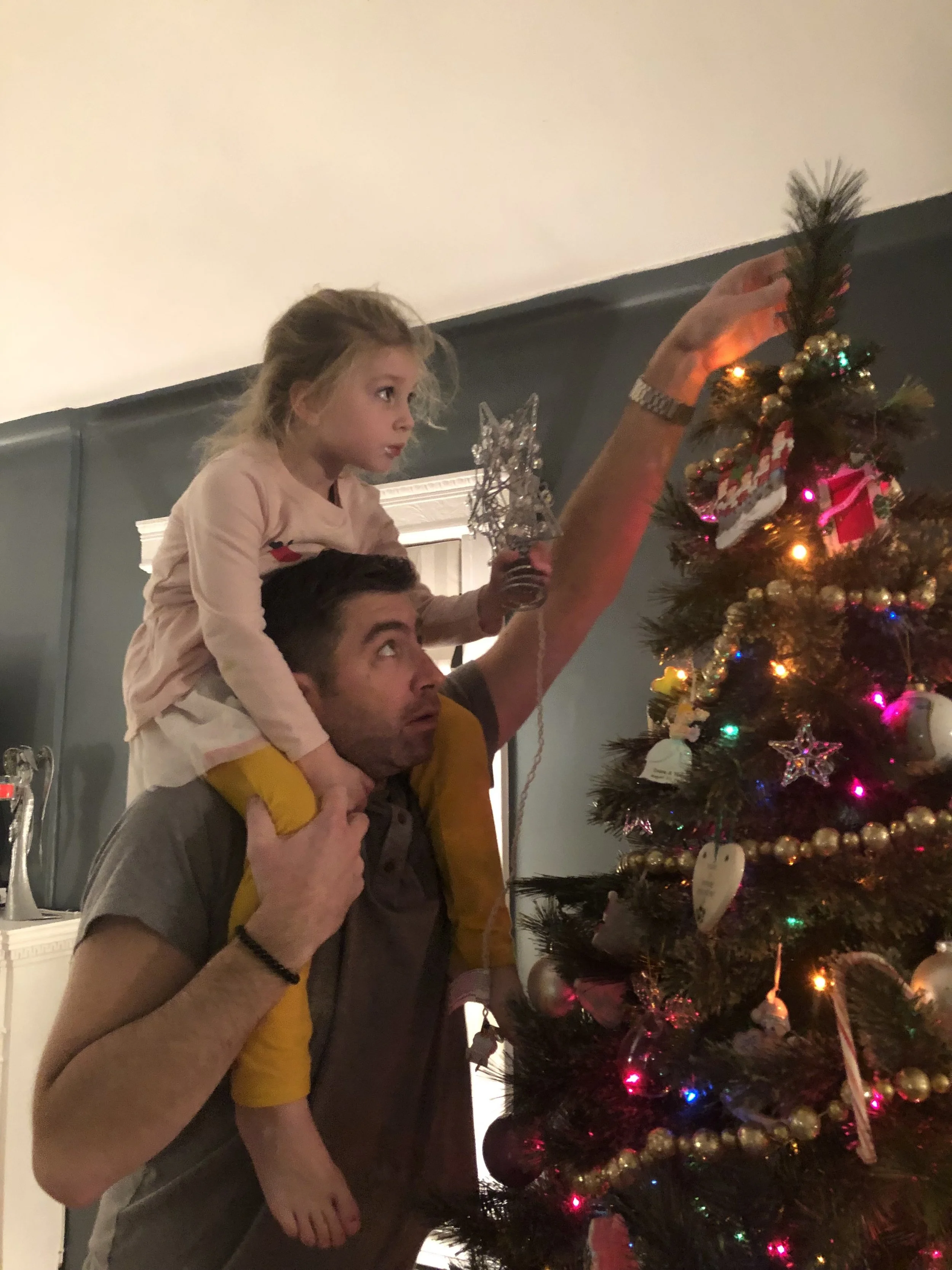 A man giving a girl a piggyback ride while they decorate a Christmas tree with ornaments and lights.