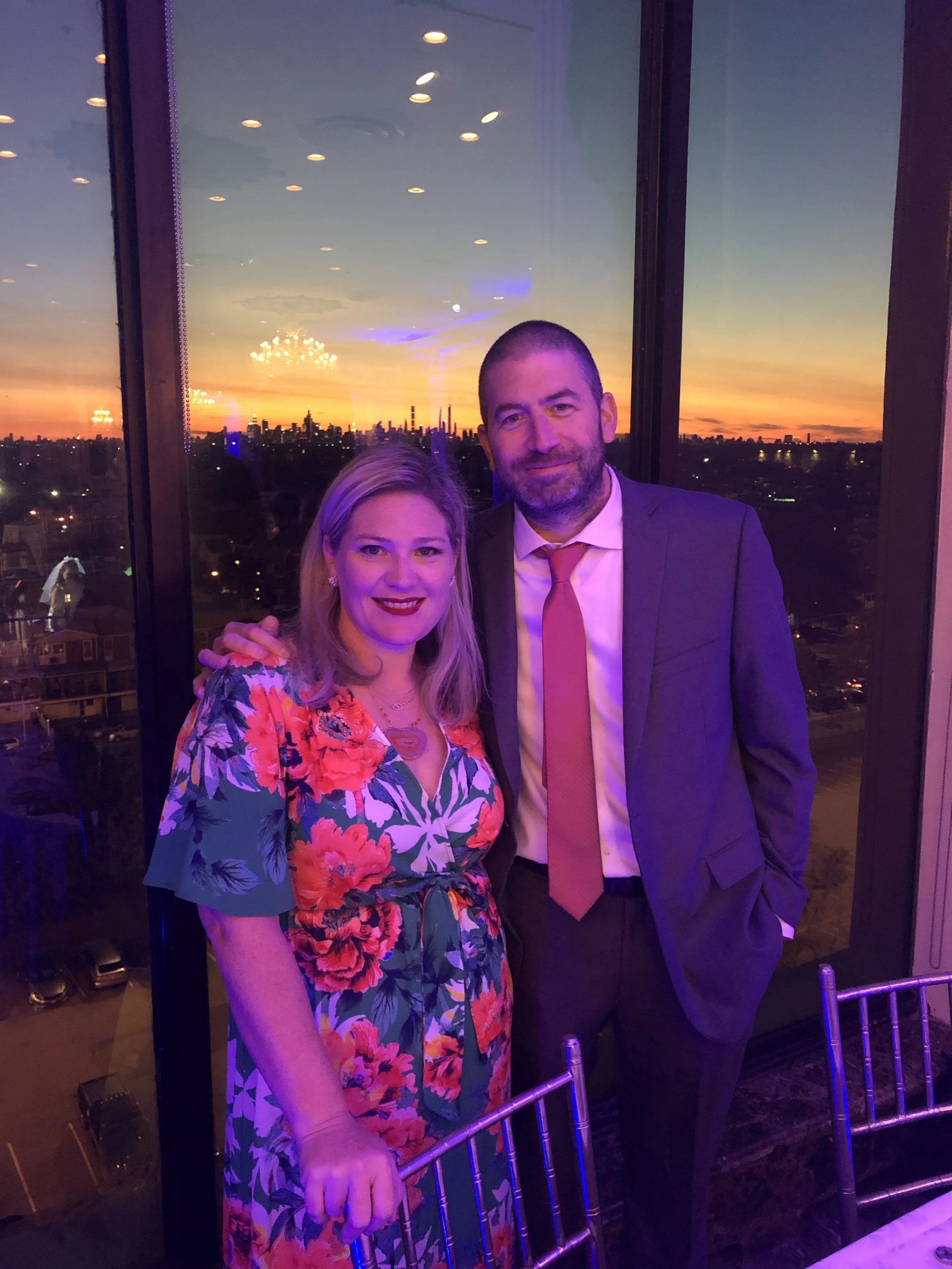 A man and woman smiling and posing together at an indoor event with a city skyline and sunset visible through large windows in the background.