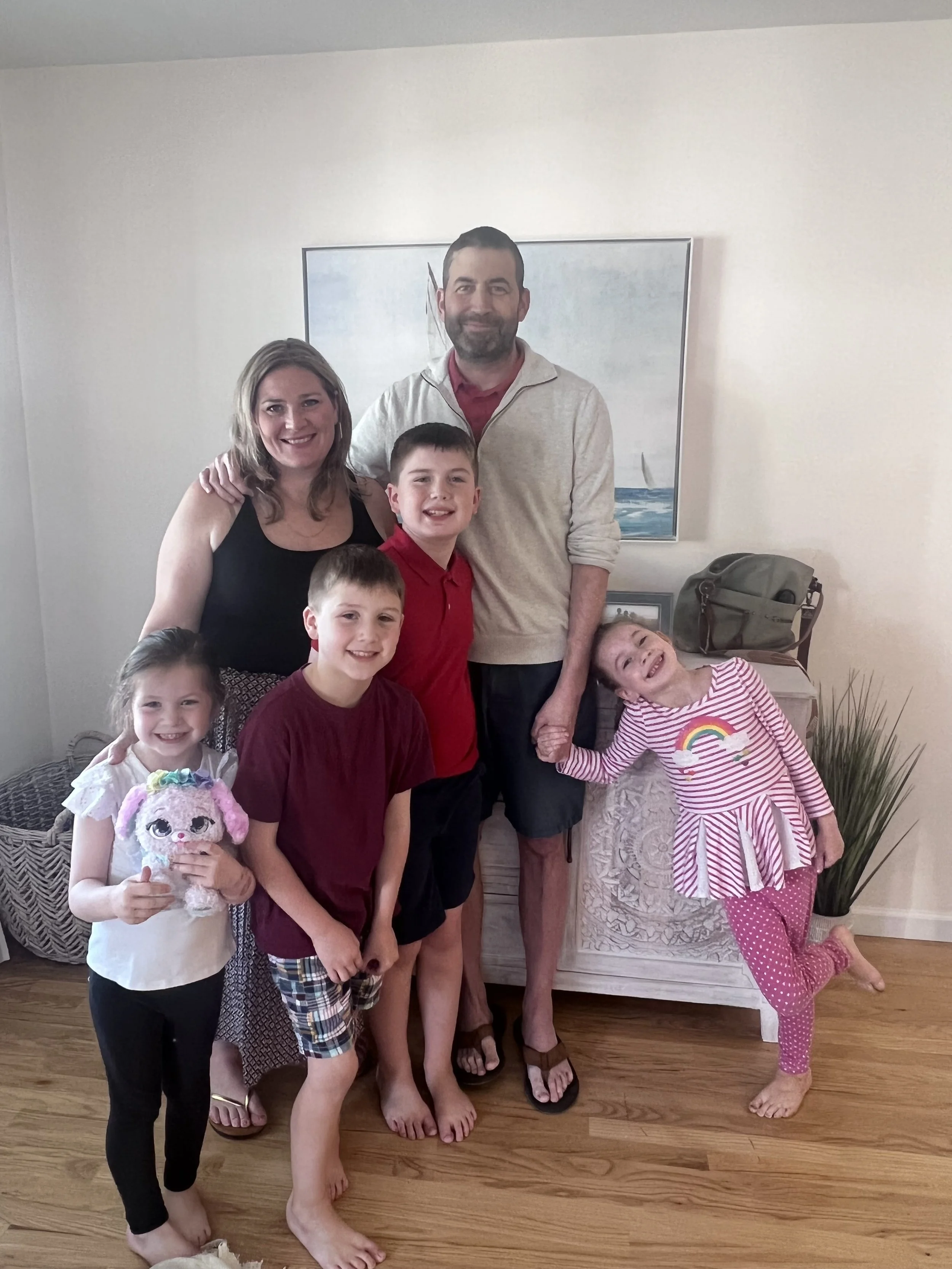 Family photo of two adults and four children standing in a living room, smiling, with a painting and a plant in the background.