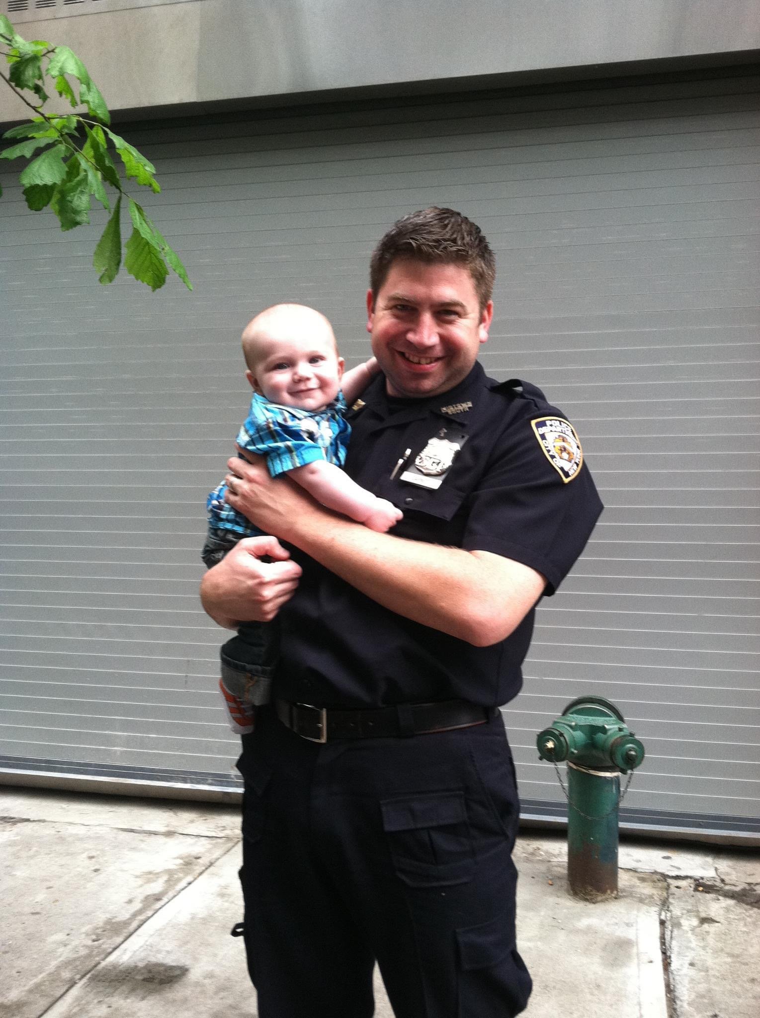 A police officer holding a smiling young child outdoors in front of a gray garage door, with a green fire hydrant nearby.