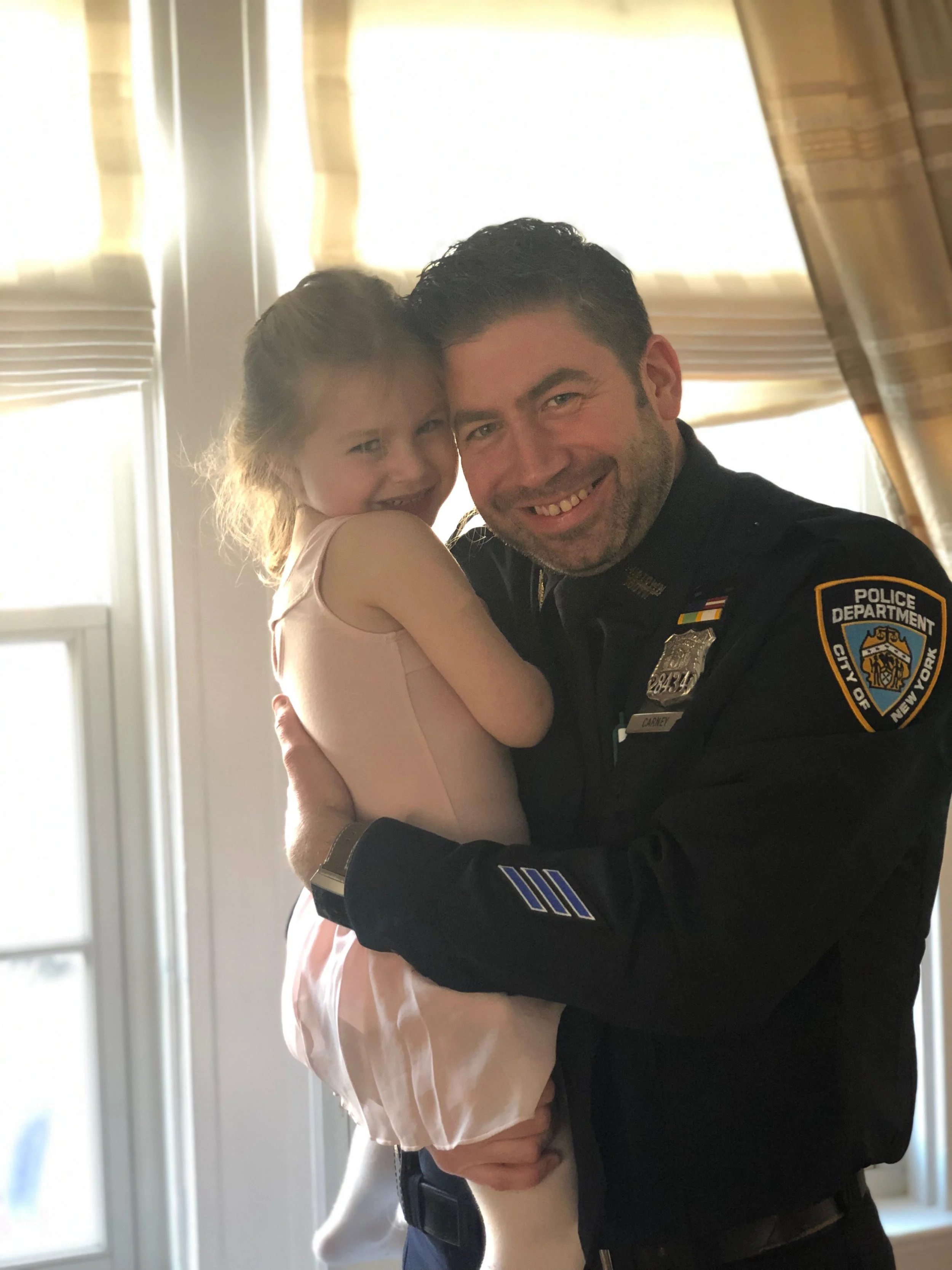 A police officer holding a young girl inside a room with curtains and a window behind them, both smiling.