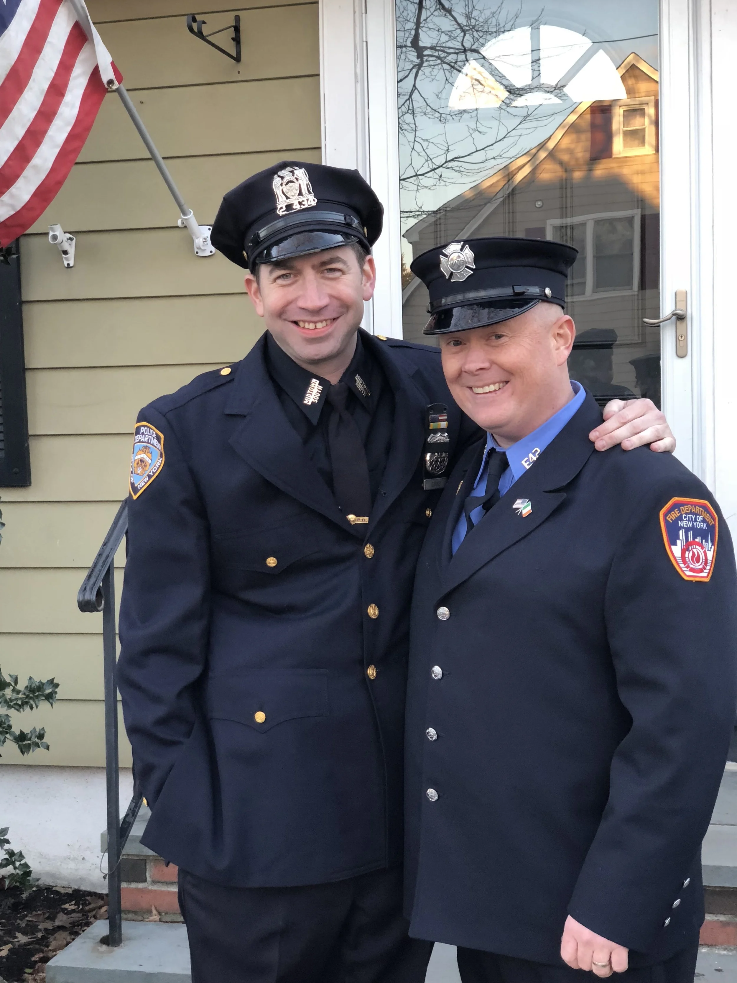 Two smiling firefighters in uniform standing outside a house, with one’s arm around the other’s shoulder. An American flag is partially visible on the left, and the house’s window and door are behind them.