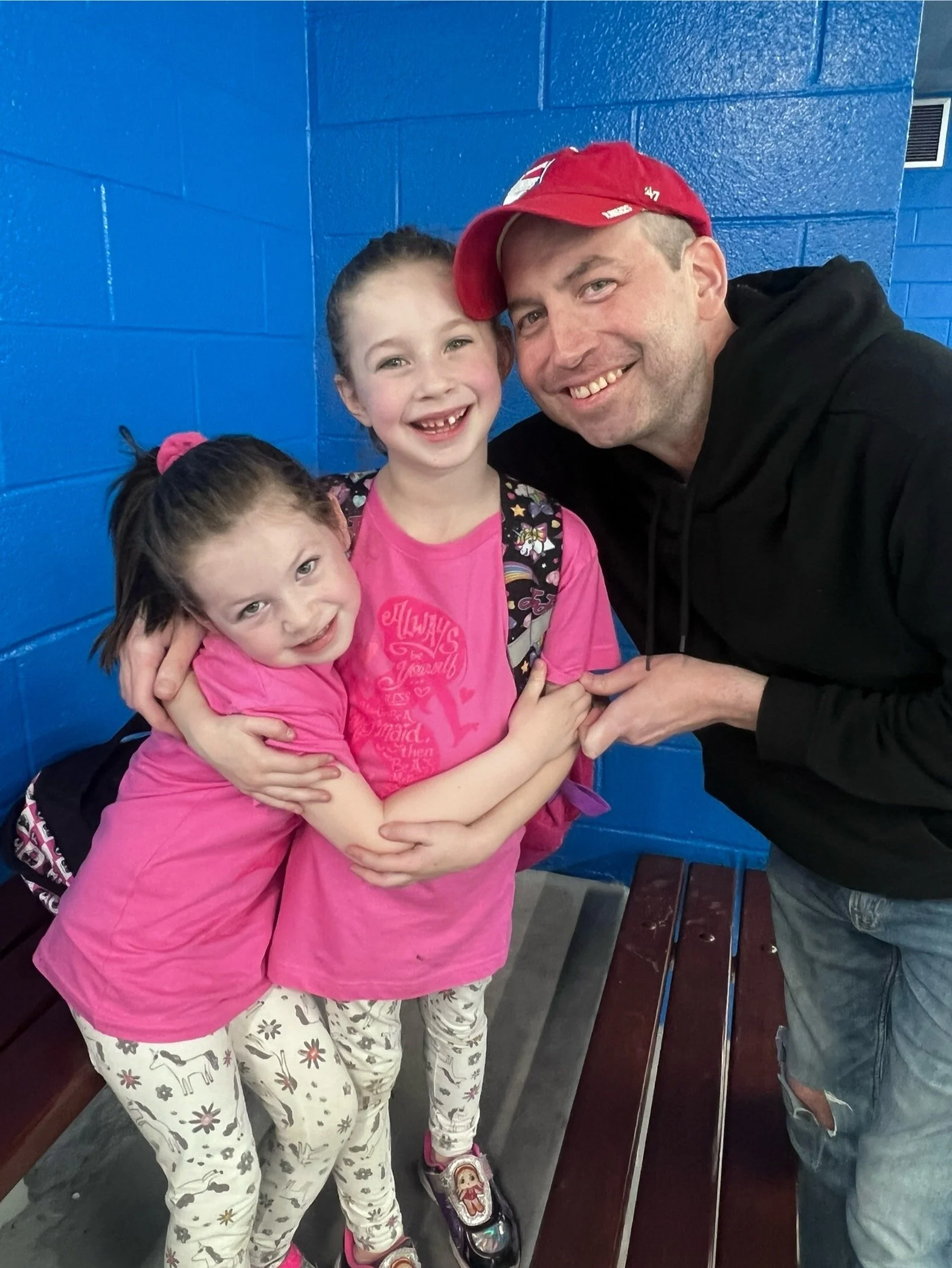 A man and two young girls smiling and hugging while standing on a bench against a vibrant blue wall. The man is wearing a red cap and a black hoodie, and the girls are wearing pink shirts and patterned leggings.
