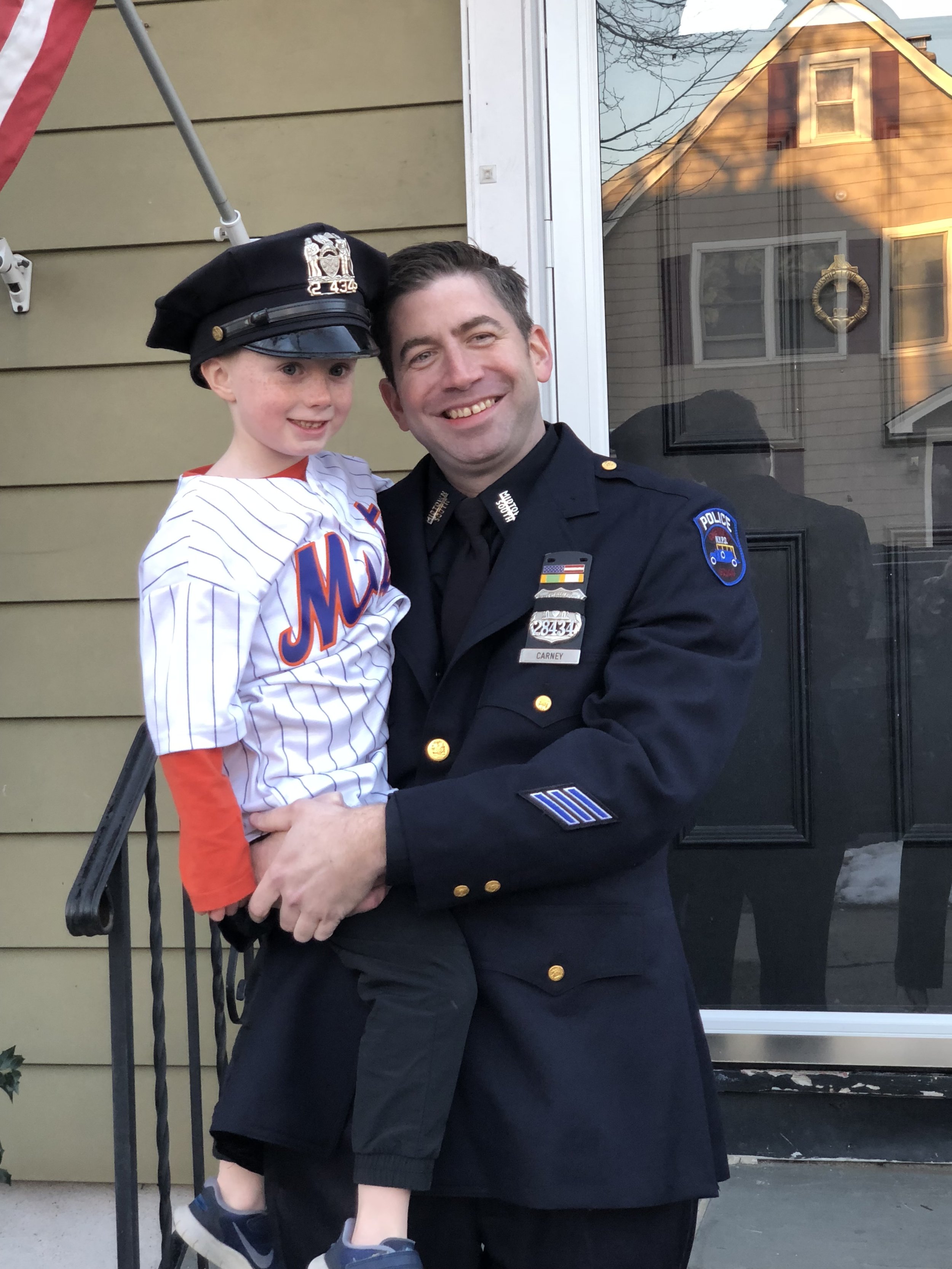A police officer holding a young boy dressed in a baseball uniform, standing outside a house with a window and door behind them.