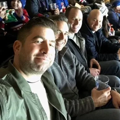 Four men sitting in seats at a sporting event or concert, smiling at the camera.