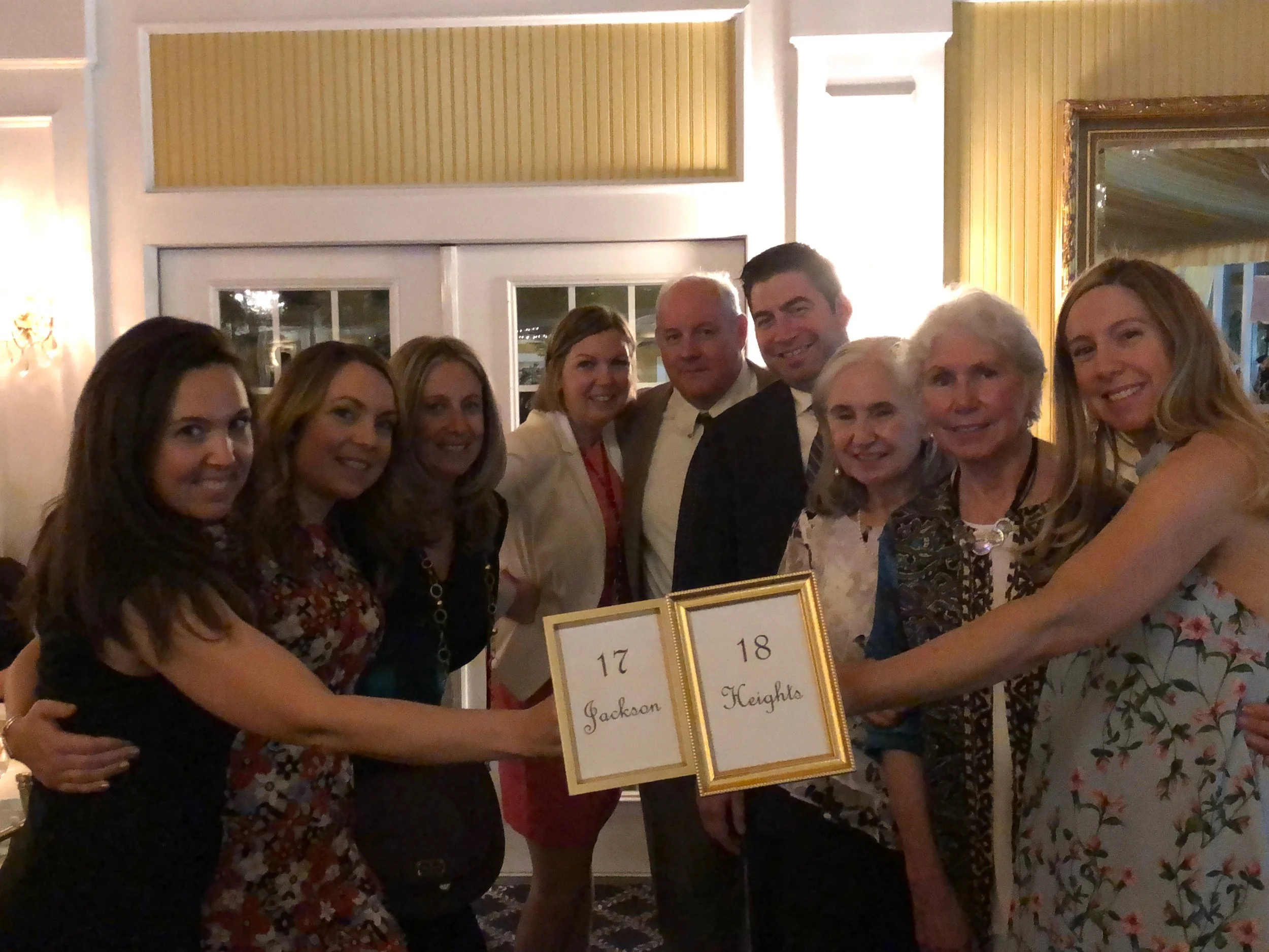 Group of nine people standing together at a table holding a framed table number that reads '17 Jackson' and '18 Heights' in a warmly lit indoor setting.