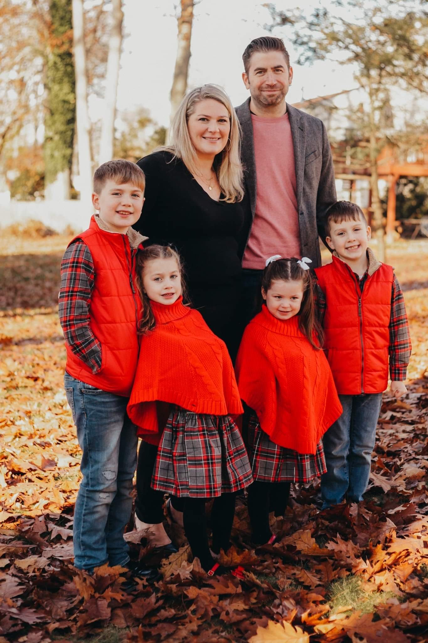 Family of six in fall outdoor setting with autumn leaves, posing for a photo. The adults are standing in the back, women in black and gray blazer, men in sweater and blazer, and children in red vests and tartan dresses and pants.