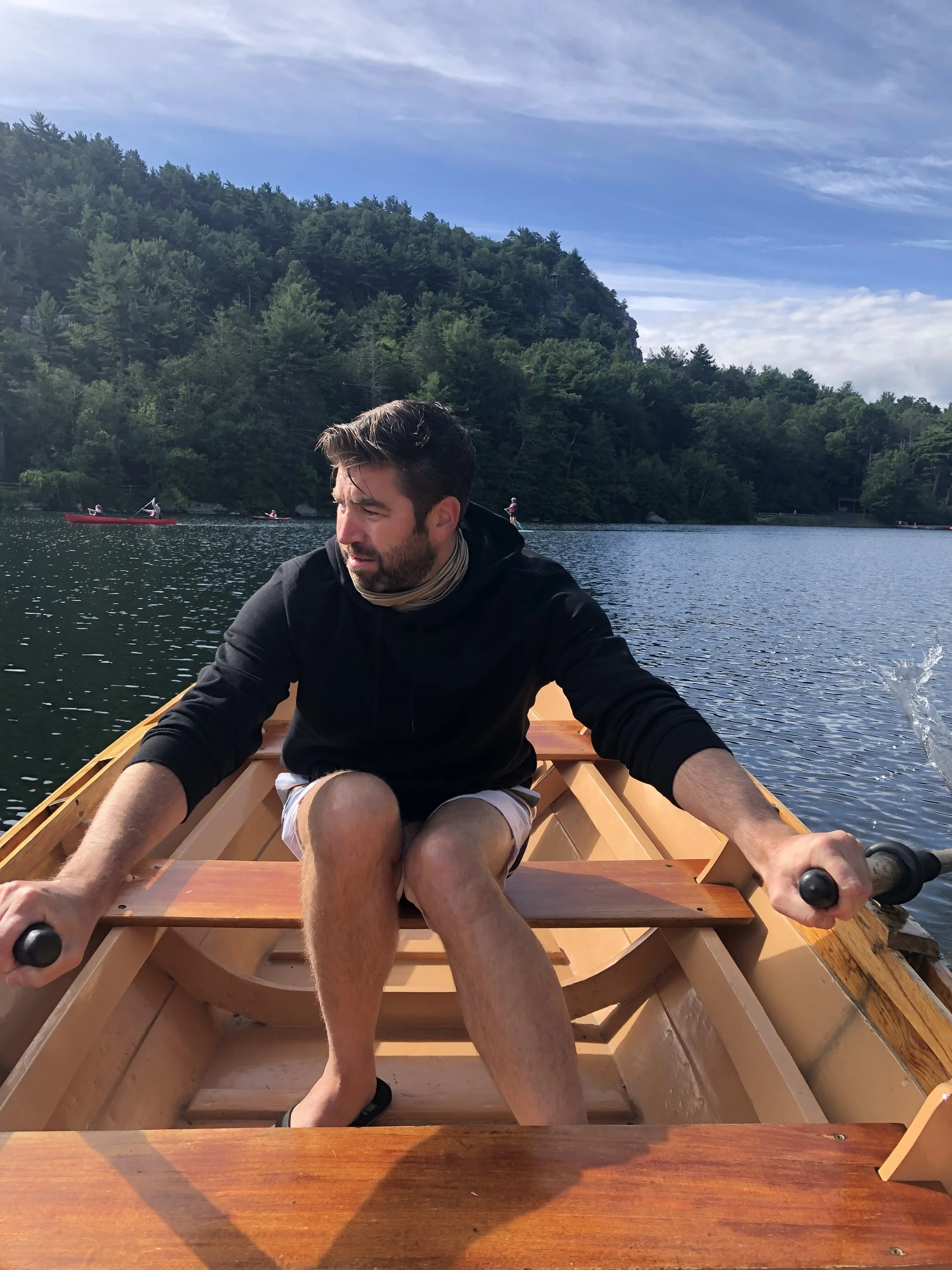 A man rowing a boat on a lake with green trees and hills in the background, under a partly cloudy sky.
