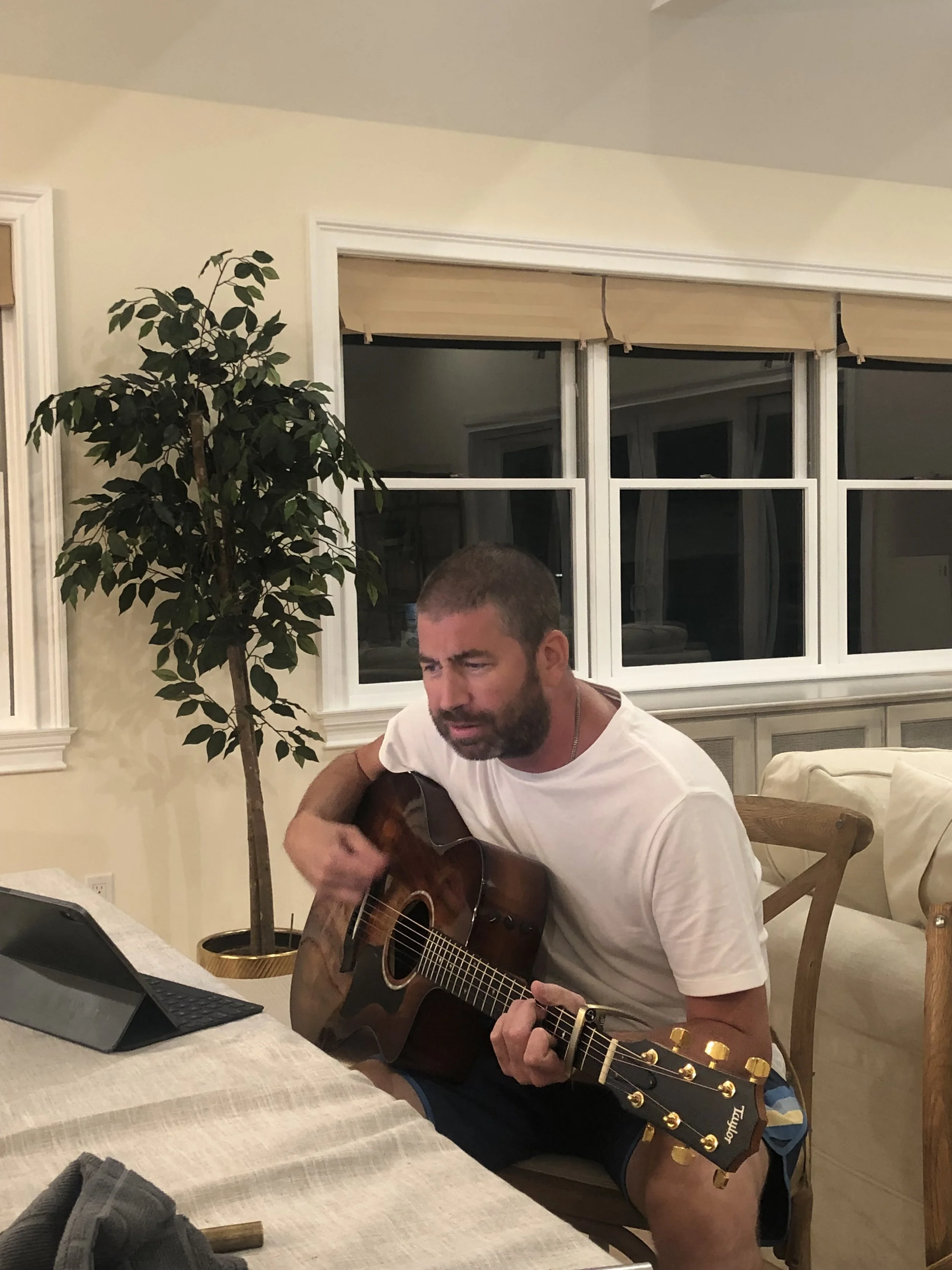 A man is sitting at a dining table playing an acoustic guitar, wearing a white t-shirt and shorts, with windows and a potted plant in the background.