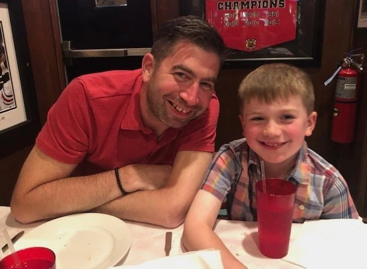 A man and a boy are sitting at a restaurant table, smiling at the camera. The man has short dark hair and a beard, wearing a red shirt. The boy has blonde hair, wearing a colorful plaid shirt with a straw in his red cup.