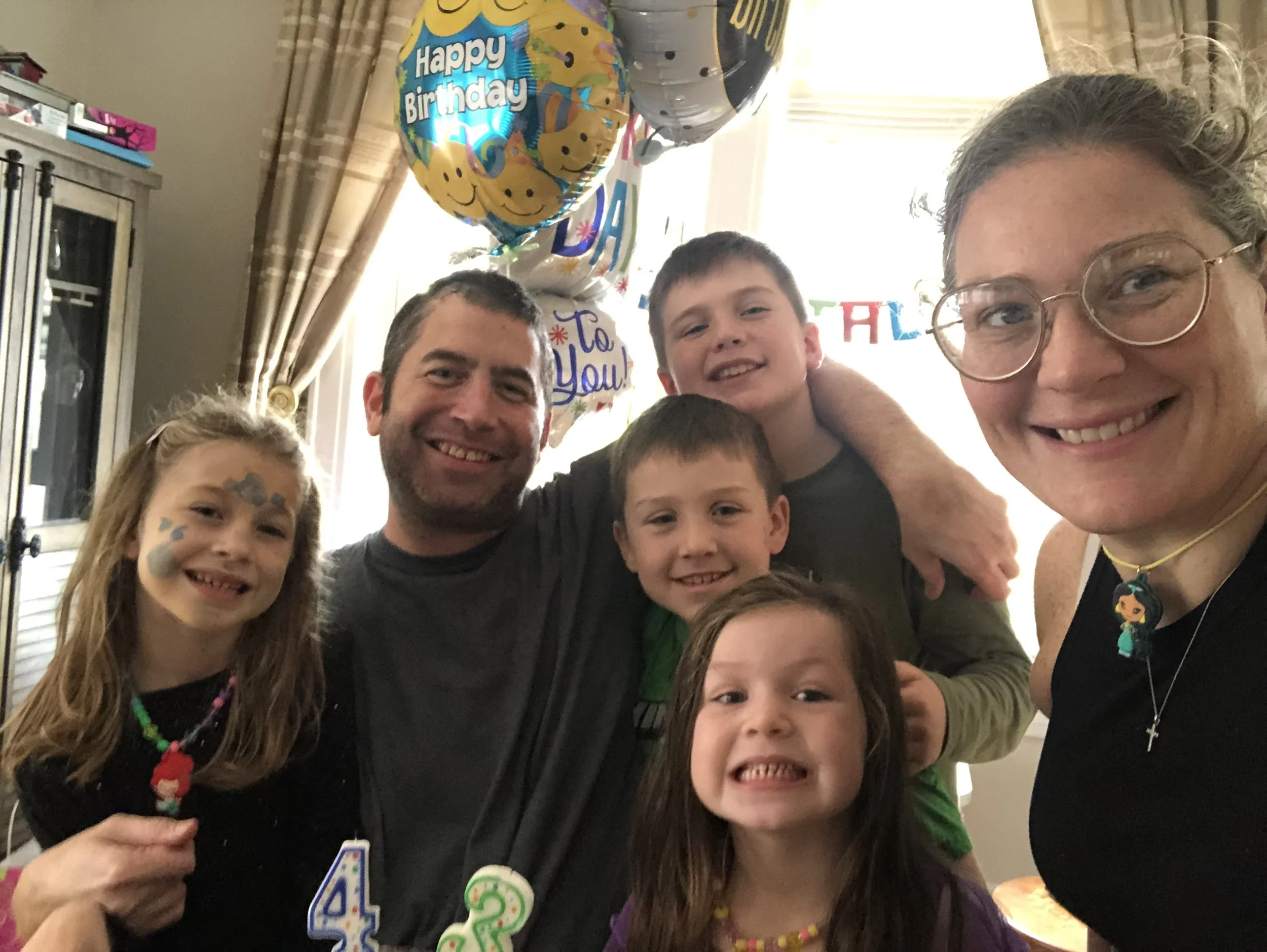 A birthday celebration with a family group of six smiling children and two adults in a room decorated with birthday balloons, including one that says "Happy Birthday" and others with messages like "To You" and "Thank You."