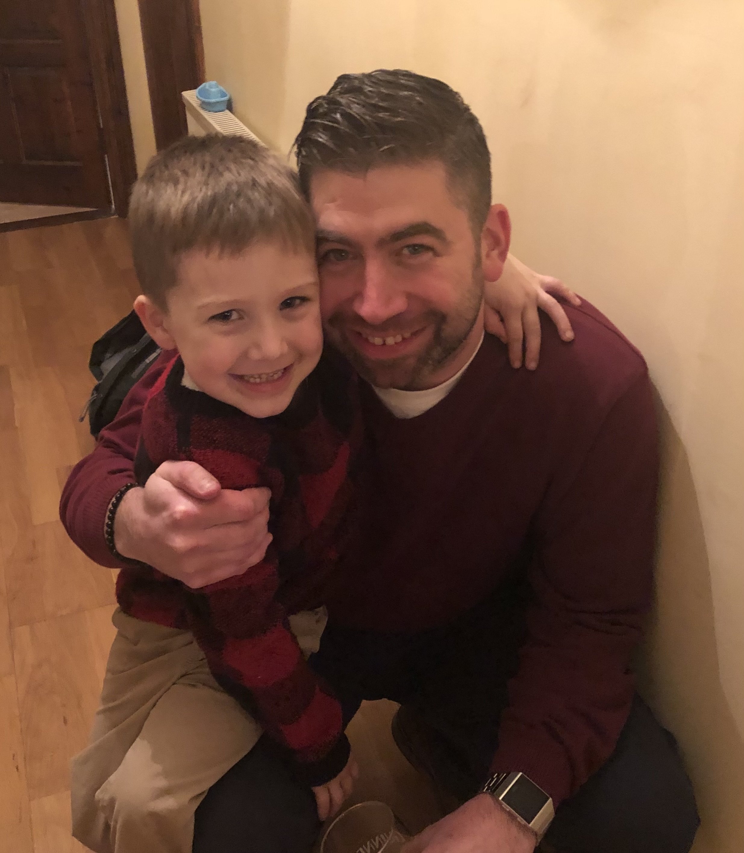 A smiling man and young boy hugging each other indoors, with the man sitting on the floor beside a wall.
