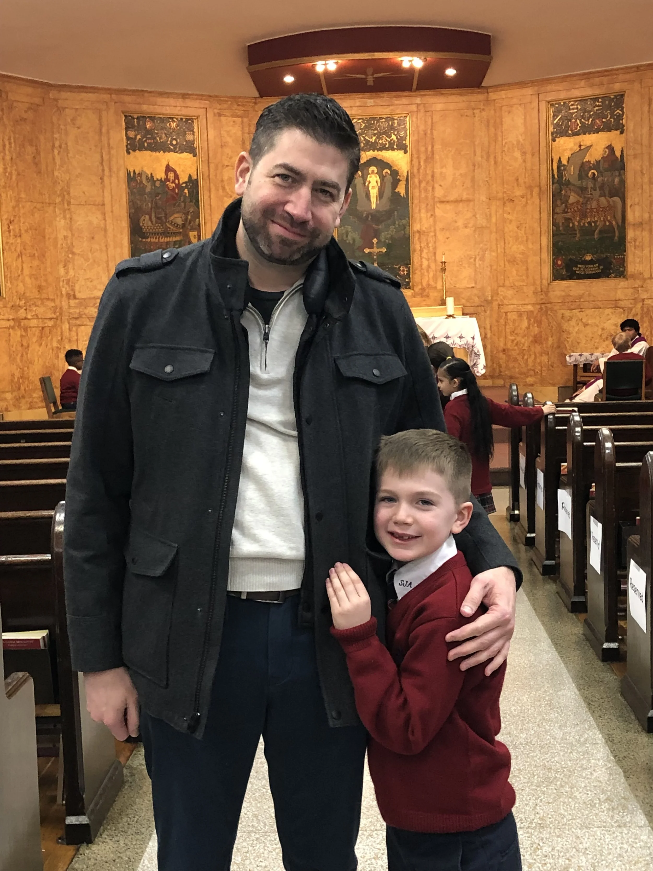A man and a young boy are standing together inside a church, smiling at the camera. The man has short dark hair and a beard, wearing a black jacket over a light-colored shirt. The boy has short light brown hair, wearing a red sweater over a white col