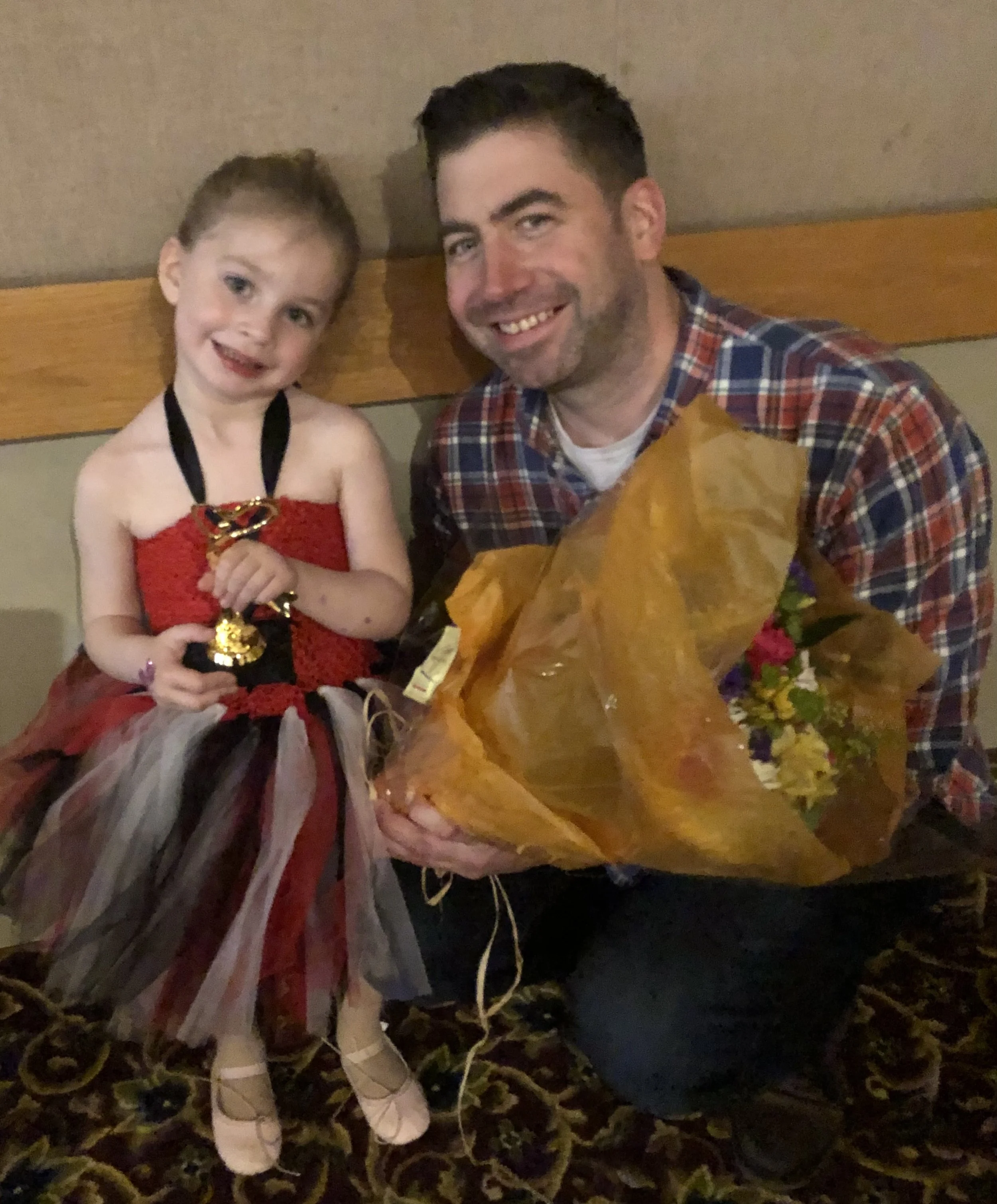 A young girl and an adult man sitting on a bench, smiling. The girl is wearing a red and black dress with a striped skirt, and the man is holding a bouquet of flowers. The girl has a medal around her neck.